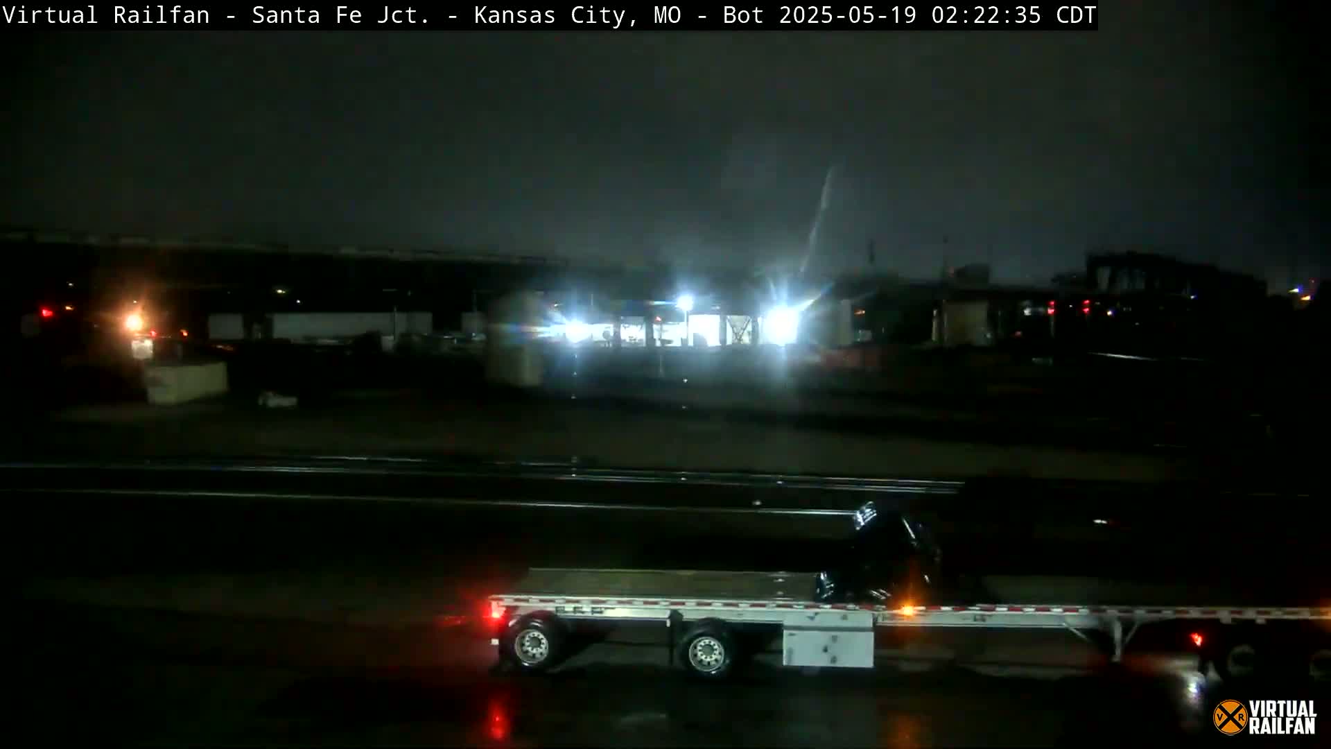 A flatbed trailer is visible at night in rainy conditions, with poorly lit buildings and structures in the background.