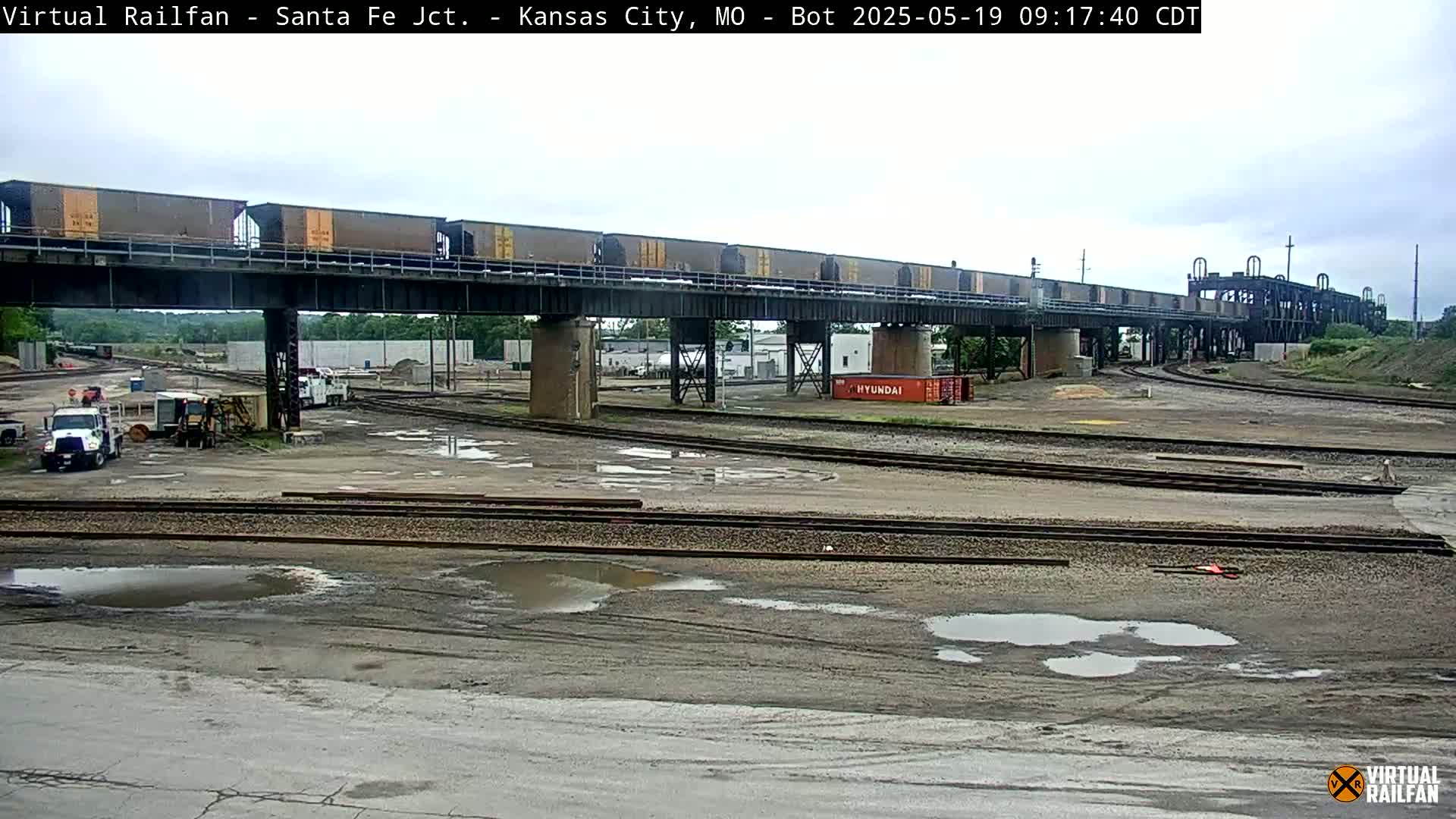 A train carrying numerous boxcars crosses a railway overpass above a rail yard with several puddles under an overcast sky.