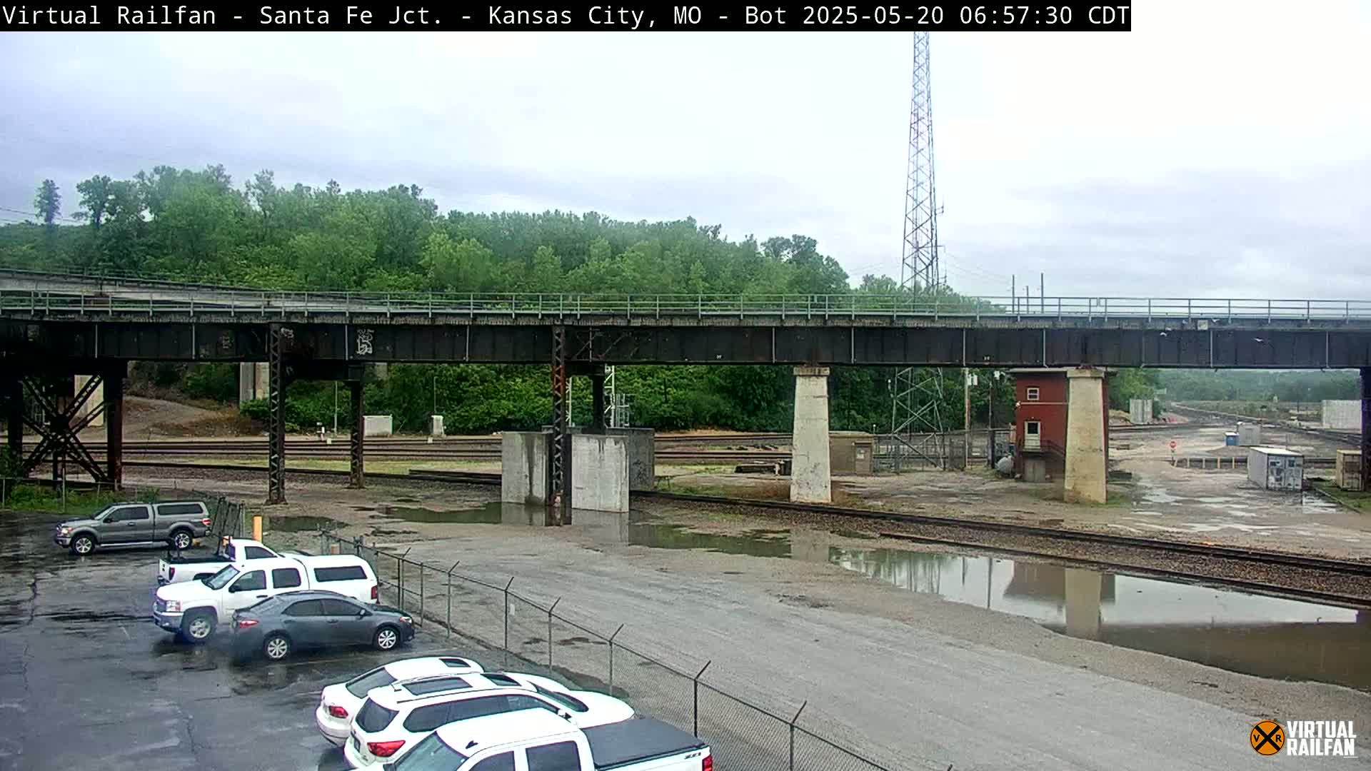 A parking lot with several trucks and cars is situated beneath a railway bridge on a cloudy day with standing water in the lot and near the tracks.