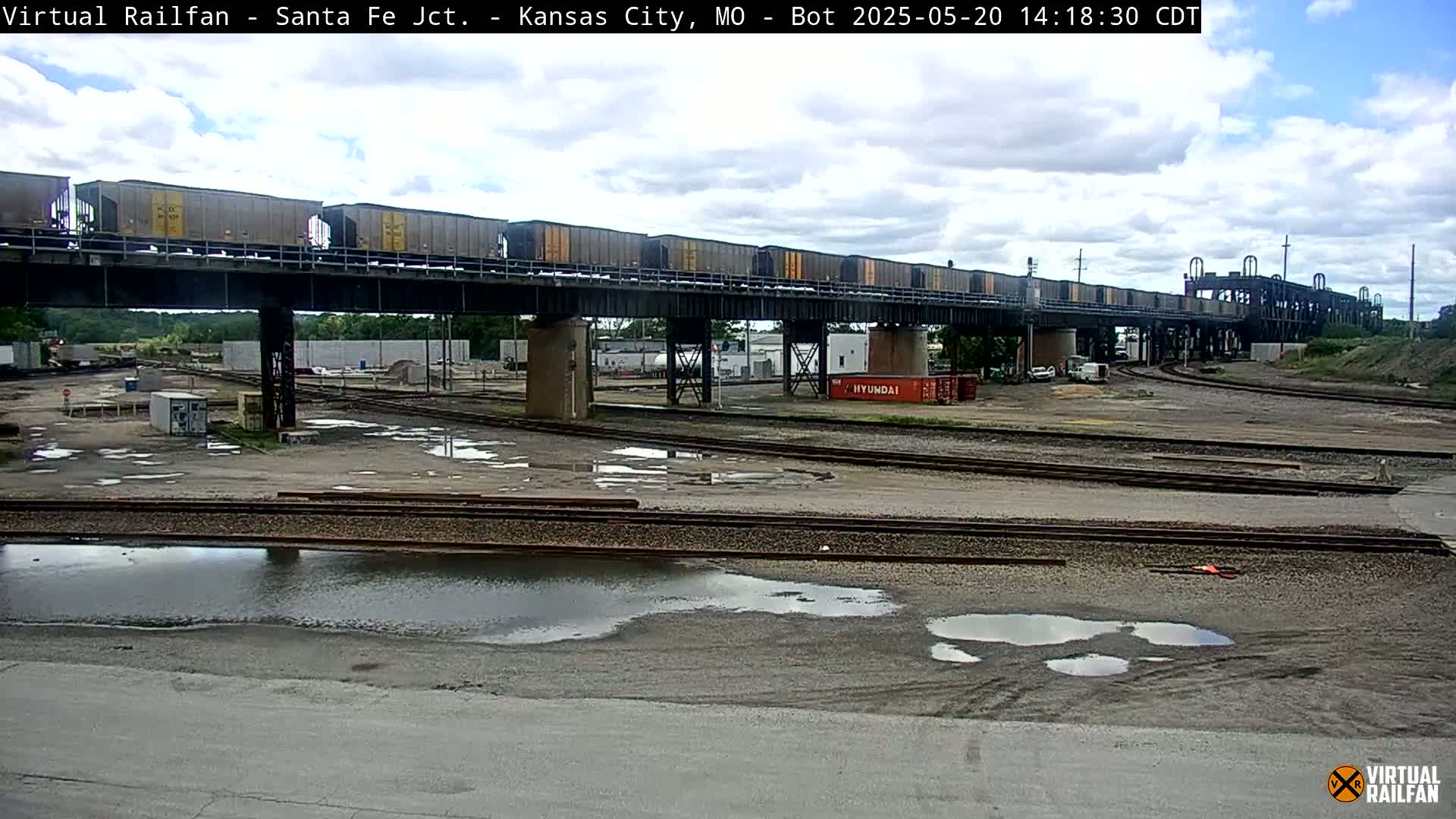 A long freight train crosses a bridge over a rail yard with puddles of water on the ground under partly cloudy skies.