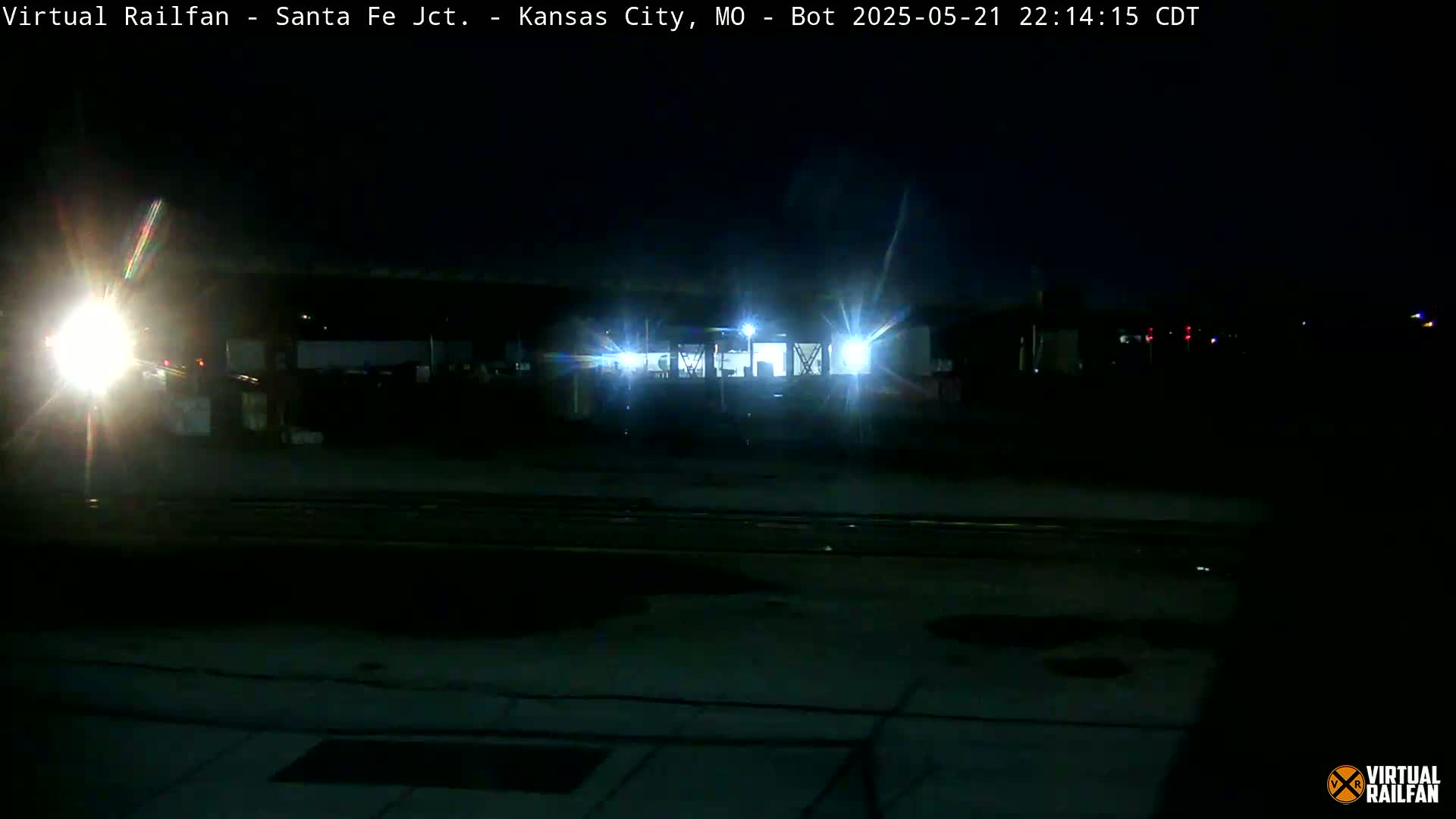 At night, bright headlights illuminate a dark area near train tracks and a building.