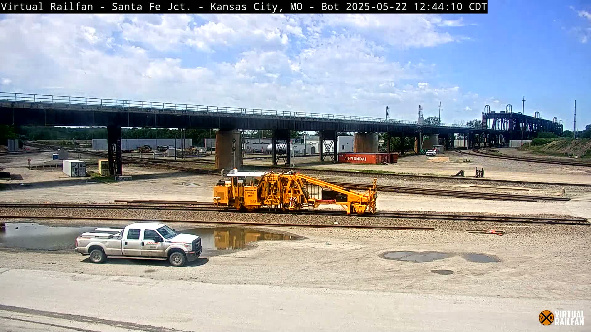A yellow rail maintenance vehicle sits on a set of tracks under a bridge on a partly cloudy day, with a white pickup truck parked nearby and several puddles in the foreground.
