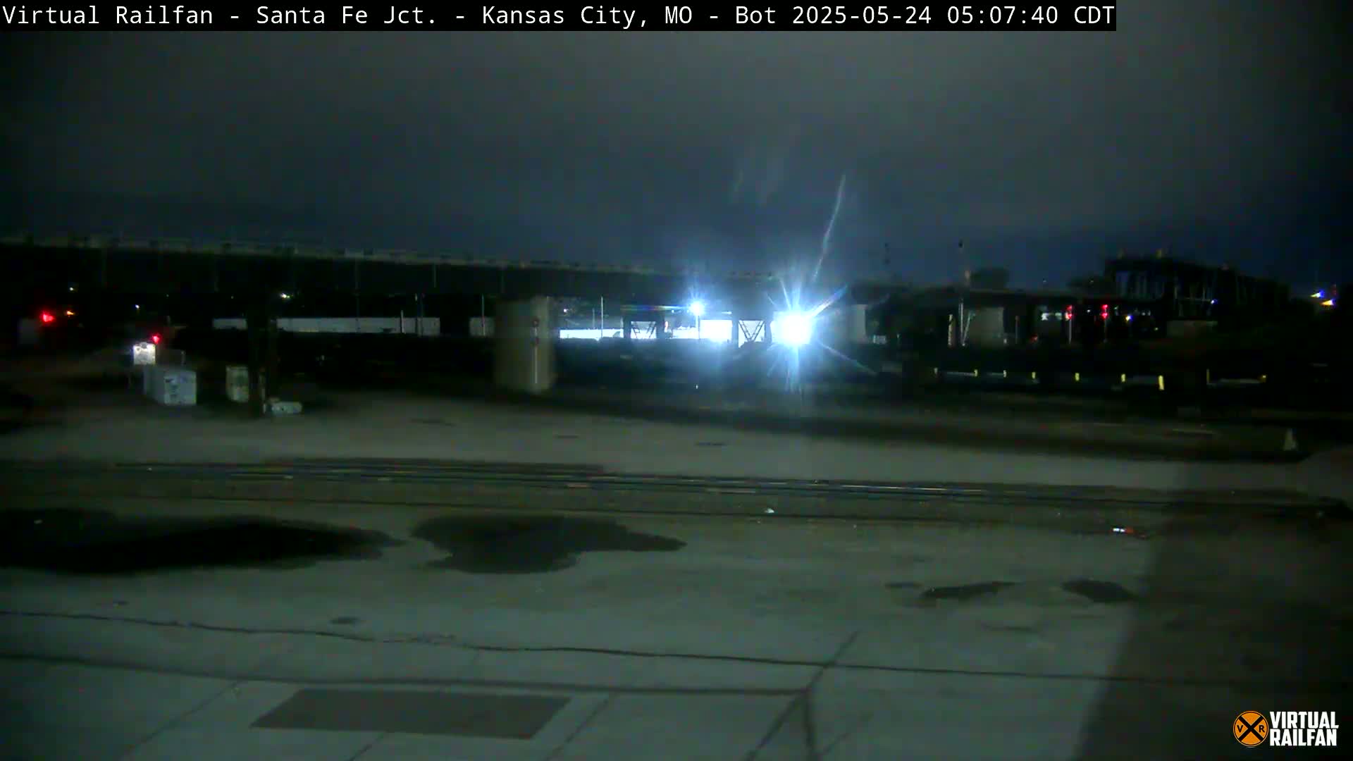 A nighttime view of a train yard with a train partially visible under a bridge.
