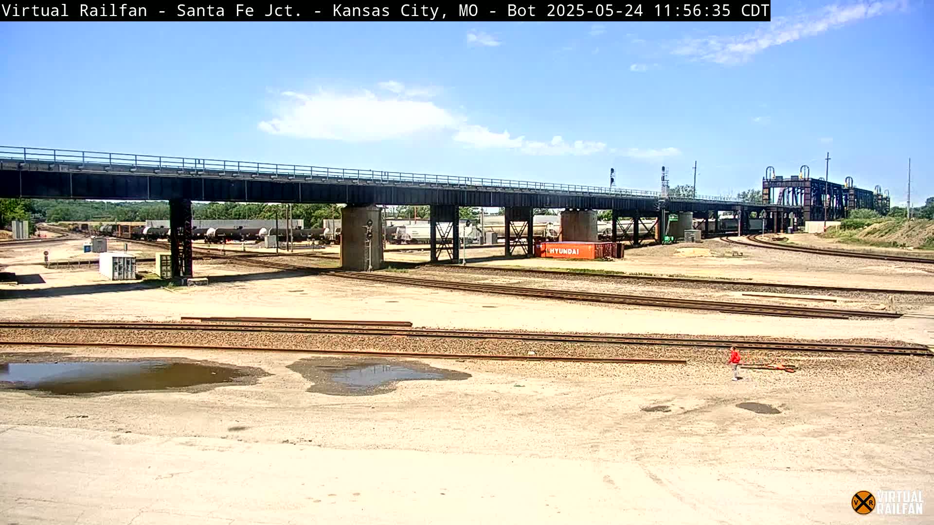 Under a mostly sunny sky, a railroad yard with numerous train cars and tracks is seen beneath a large bridge.
