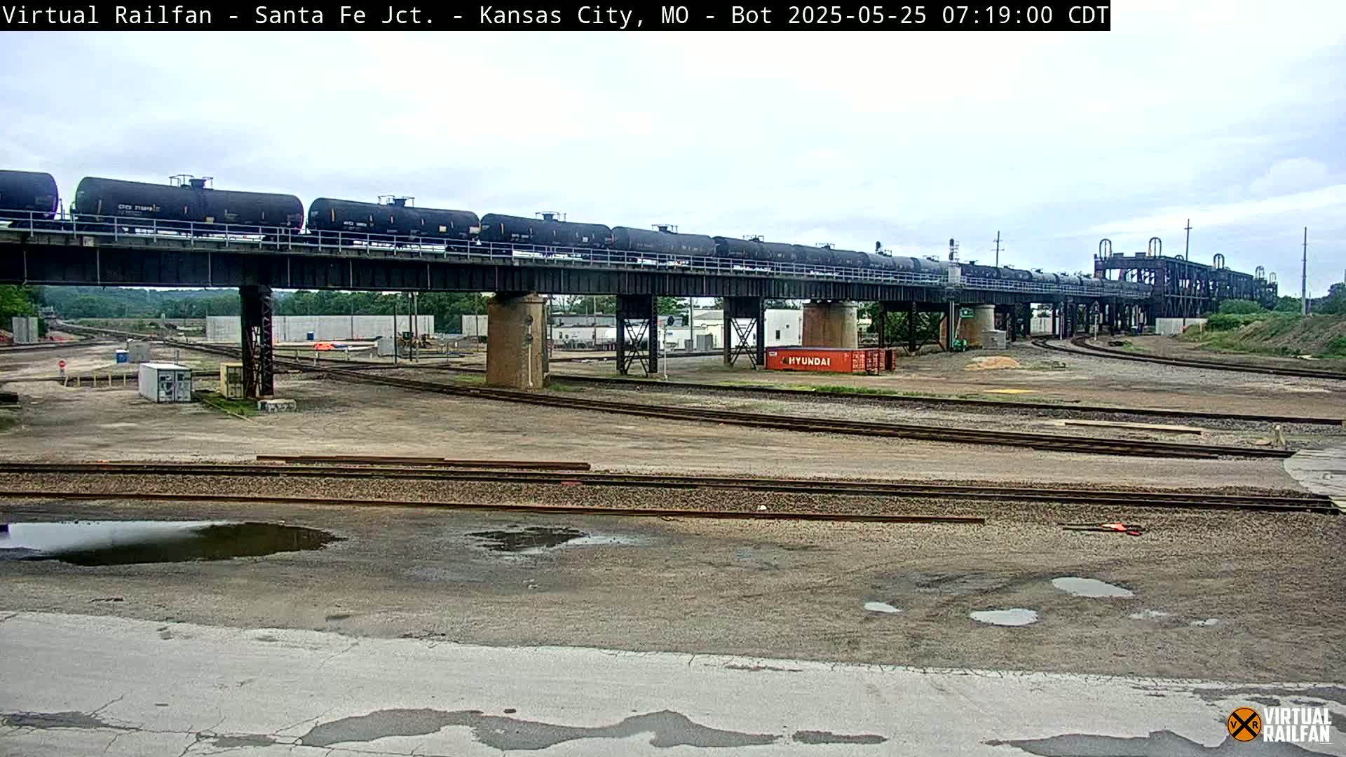 A long train of tanker cars crosses a railway bridge over a sprawling rail yard on an overcast day.
