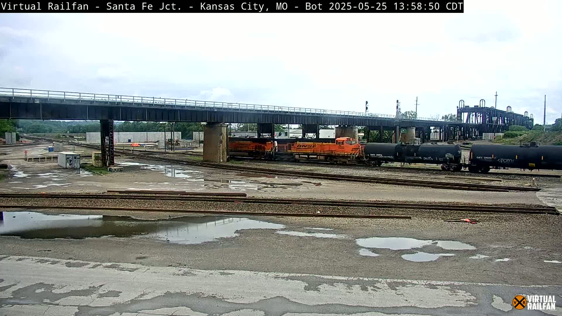 An orange and black freight train sits under a bridge in a railyard on an overcast day with puddles on the ground.