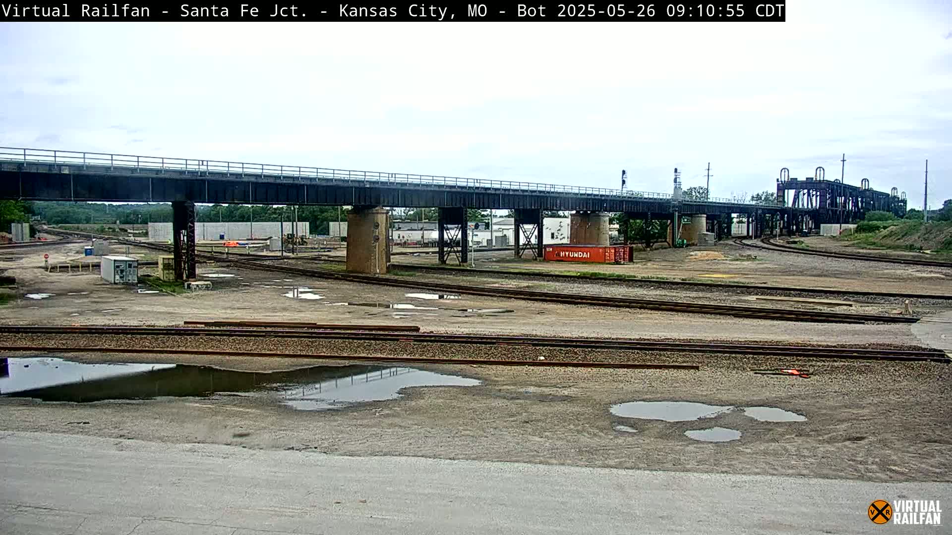 A railway yard with a large bridge overhead, several puddles on the ground, and overcast skies.
