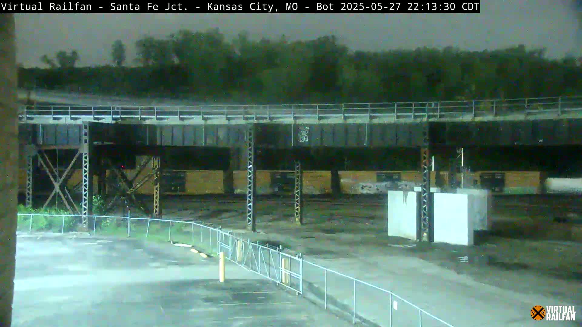 A nighttime view of a rail yard under a bridge, with a chain-link fence separating the yard from a paved area.