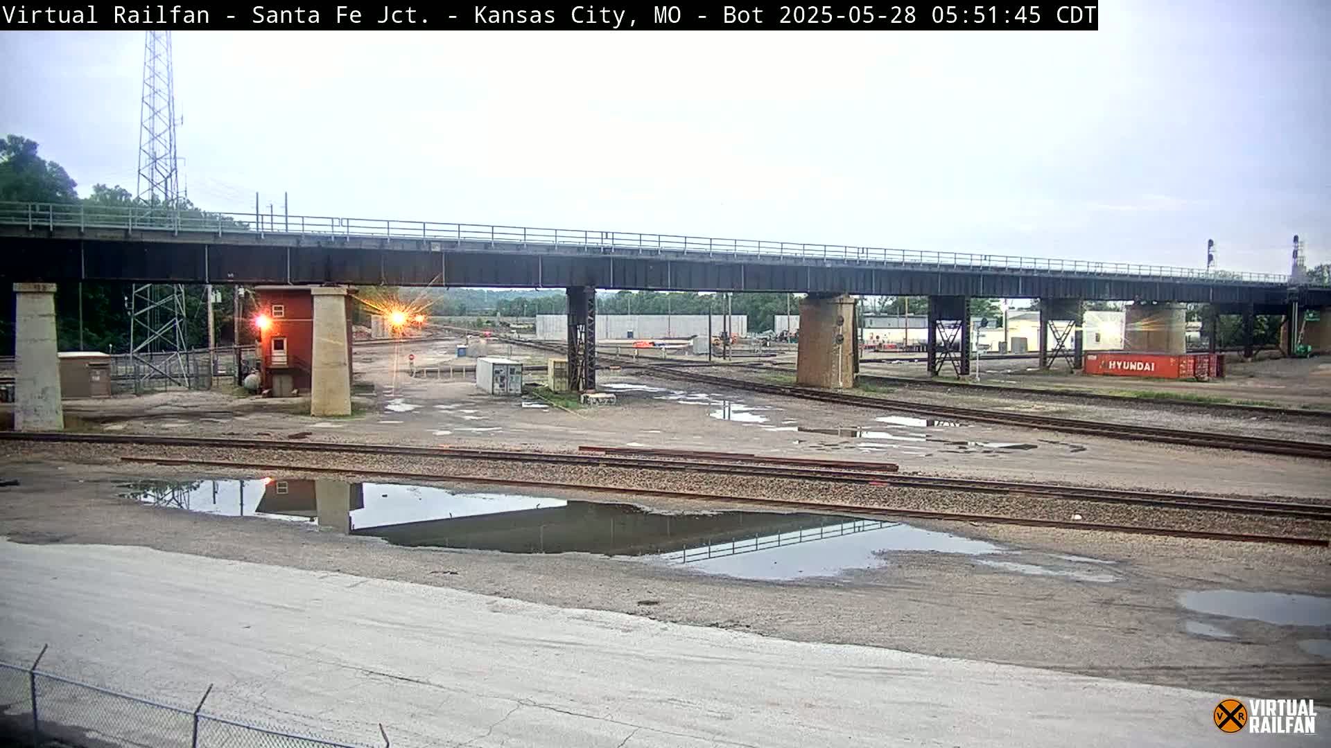 A railroad yard with a bridge overhead, under an overcast sky with puddles on the ground.