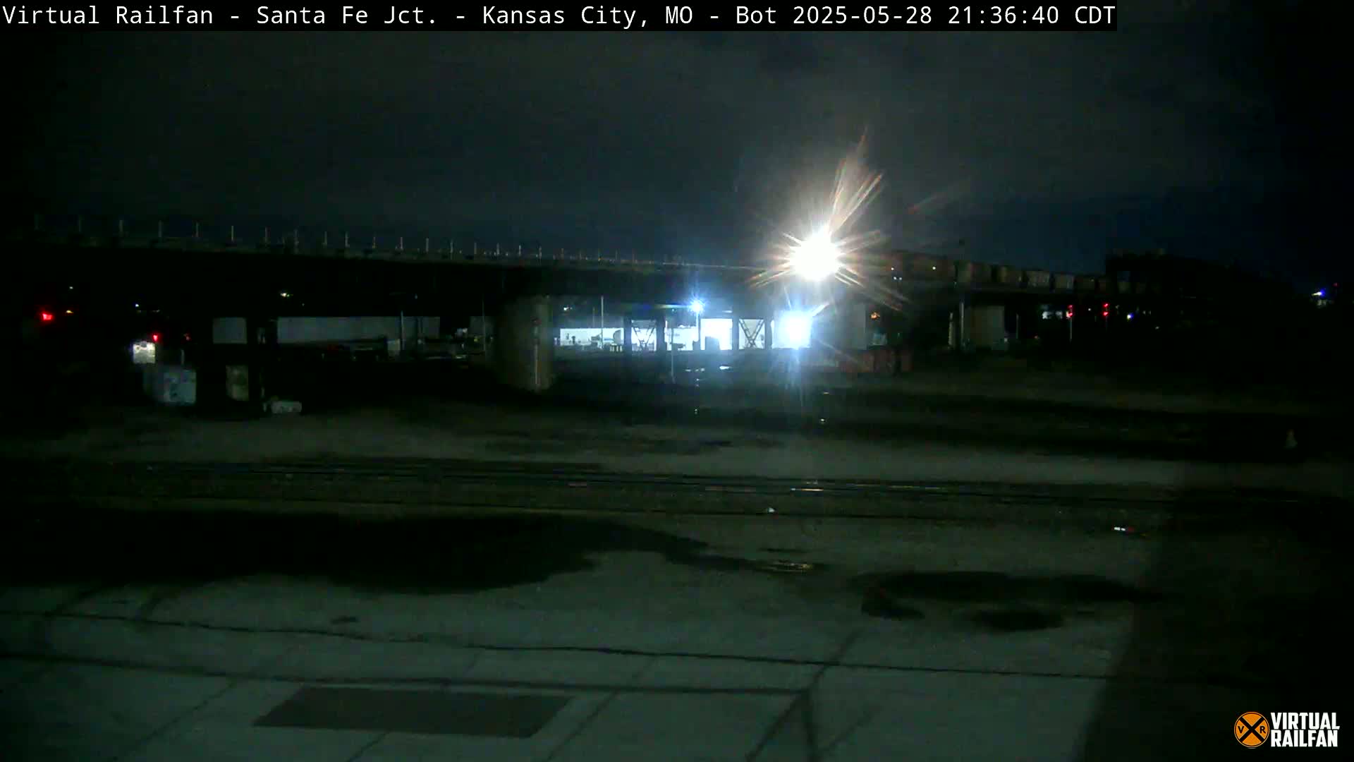 A nighttime view of a dimly lit train yard under a bridge, with a bright light source in the distance.