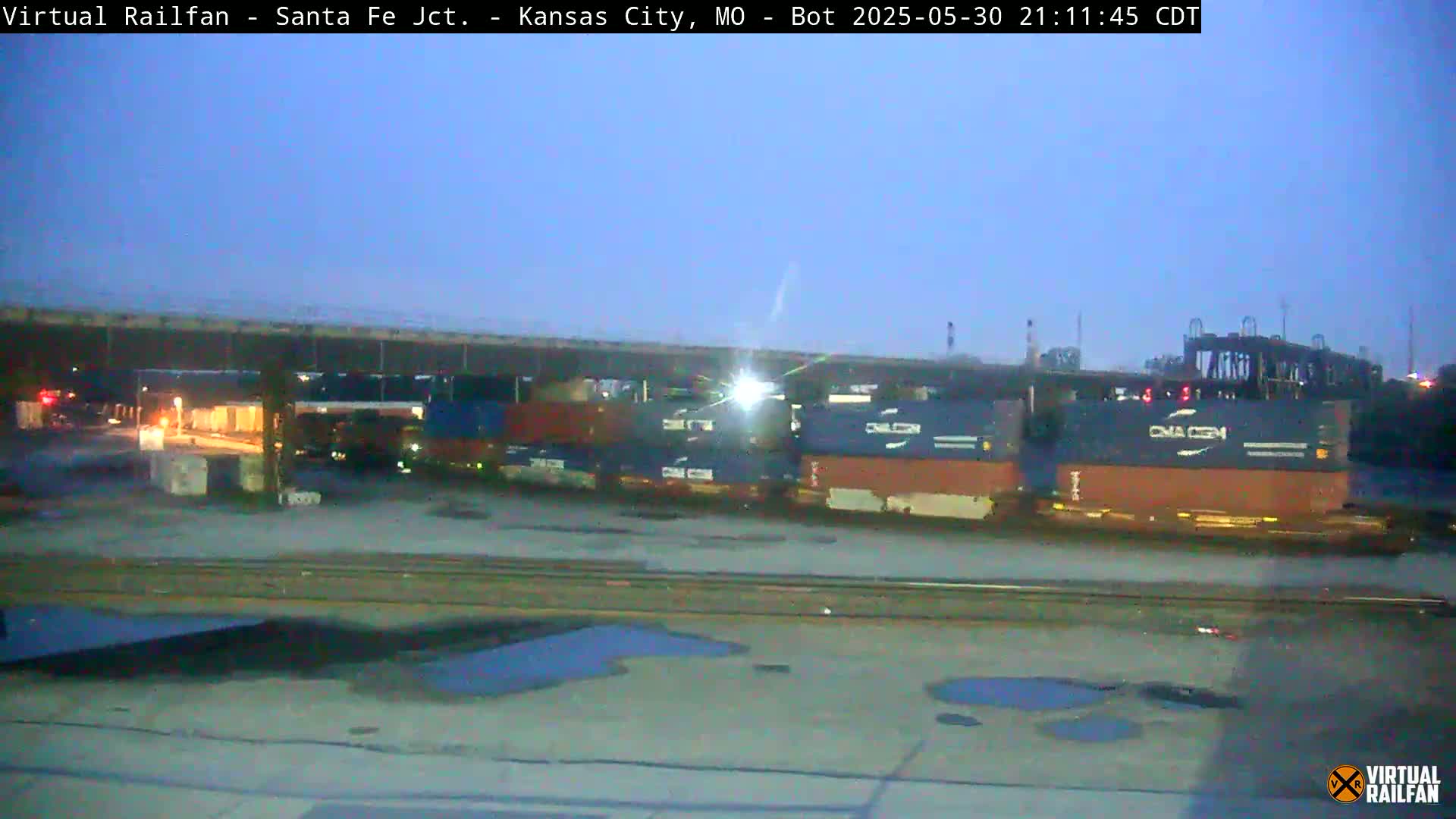 Under a twilight sky, a train carrying multiple shipping containers sits stationary near an overpass.