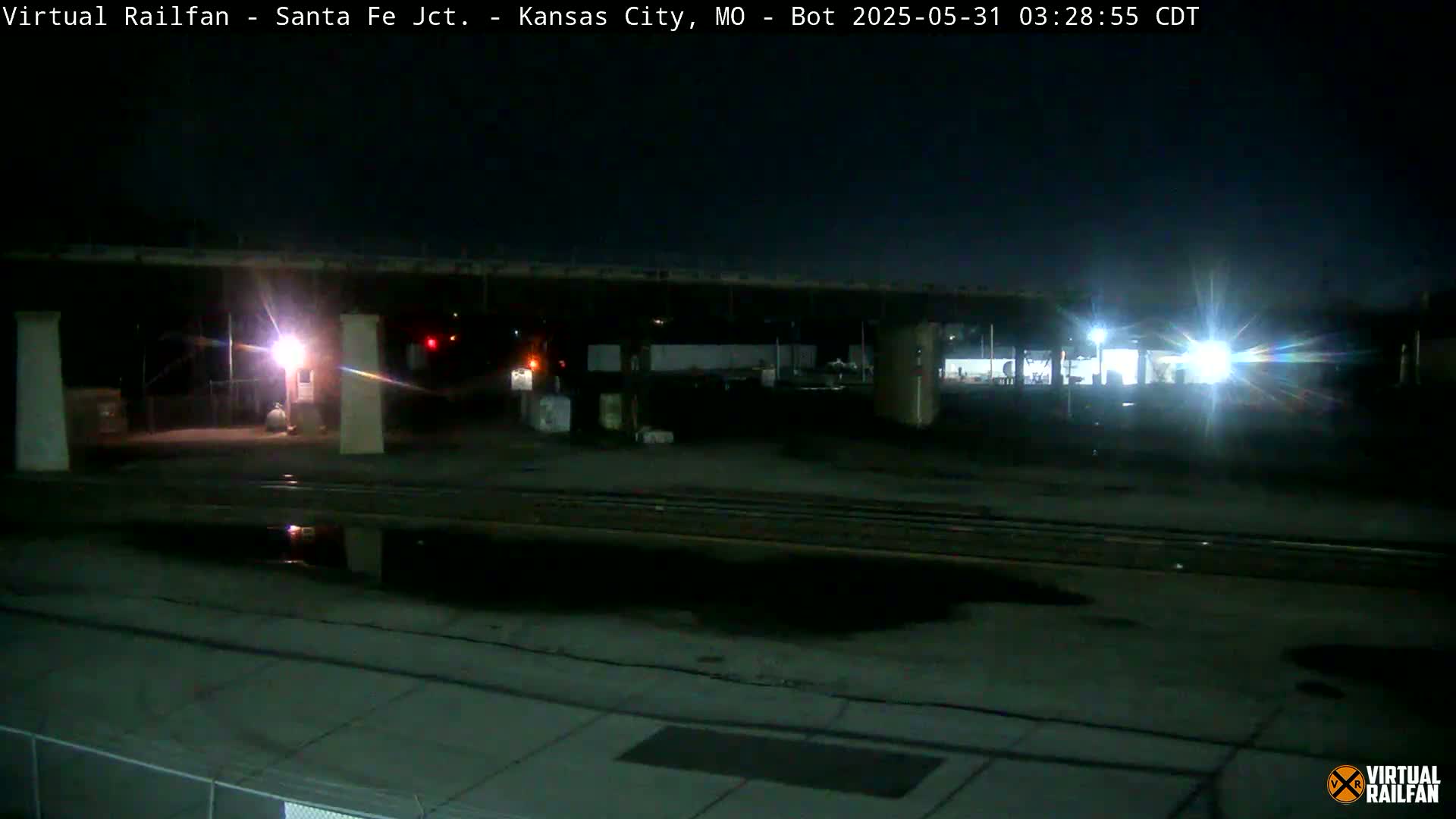 A nighttime view of a rail yard with train tracks, a roadway, and some structures under a mostly clear sky.