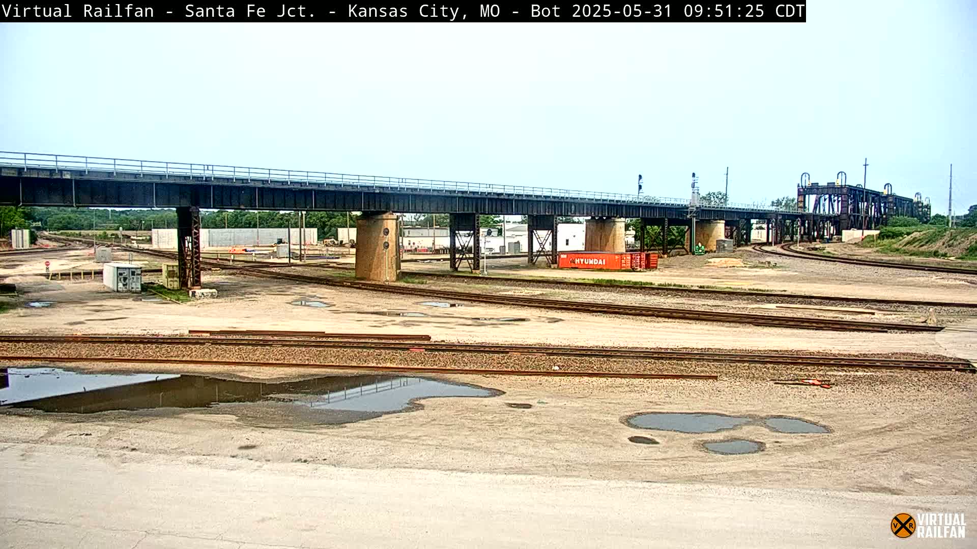 A railway yard with multiple tracks, a large bridge overhead, and several puddles of water on the ground under a mostly clear sky.
