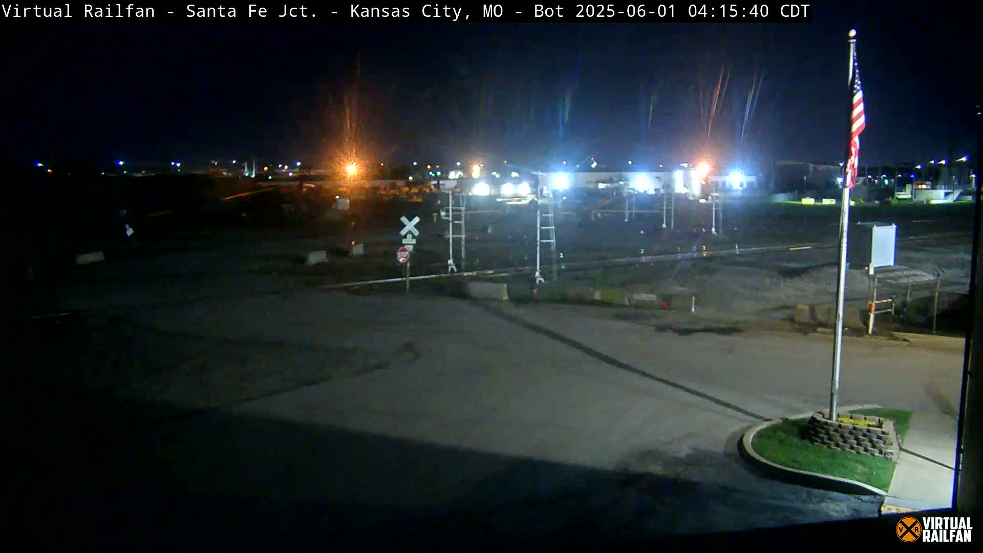 A nighttime view of a largely empty railroad crossing with a flagpole and an American flag, under a dark, clear sky.