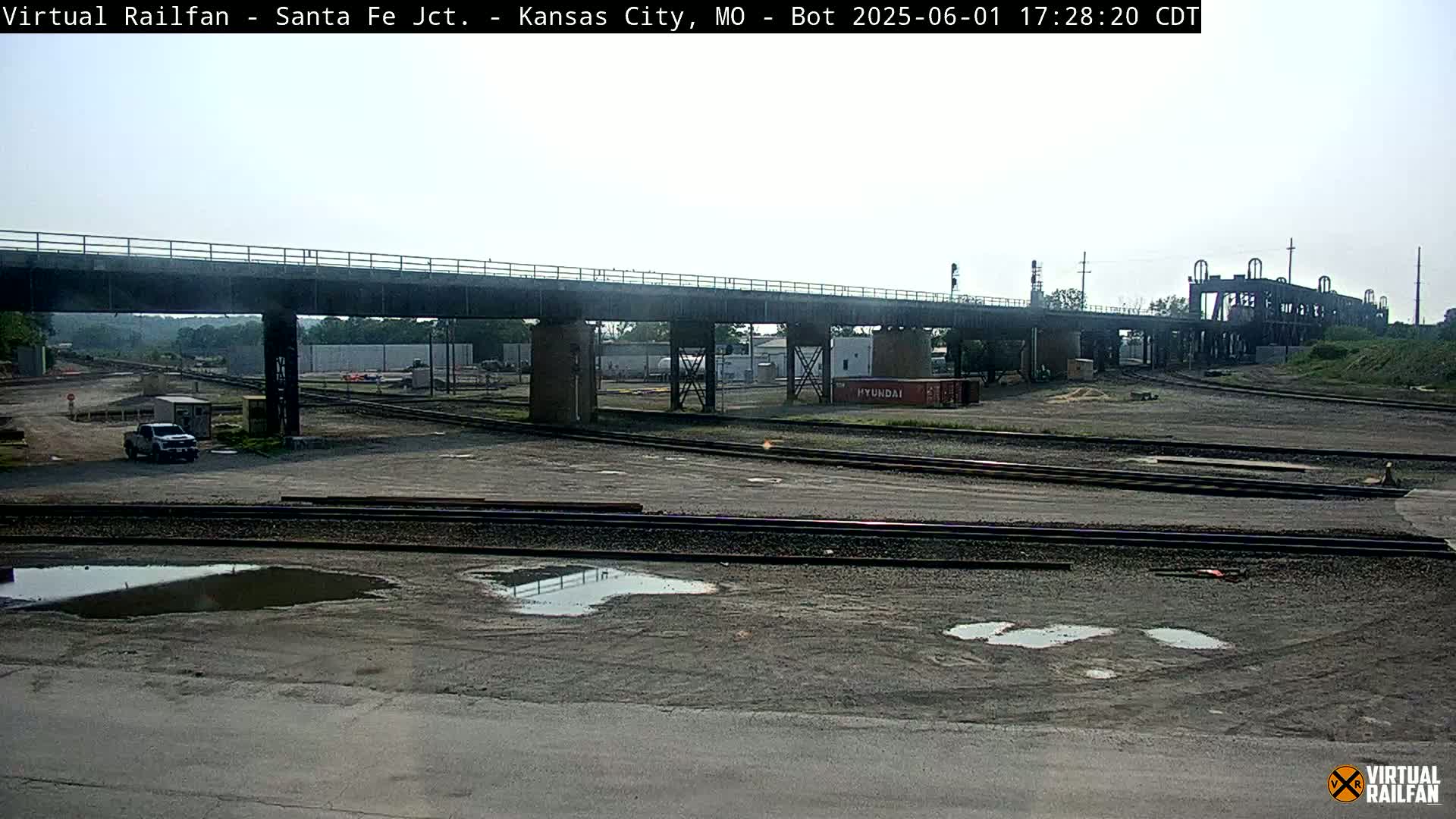 A railway yard with several train tracks, a bridge overhead, and a few puddles on the ground under a mostly clear sky.