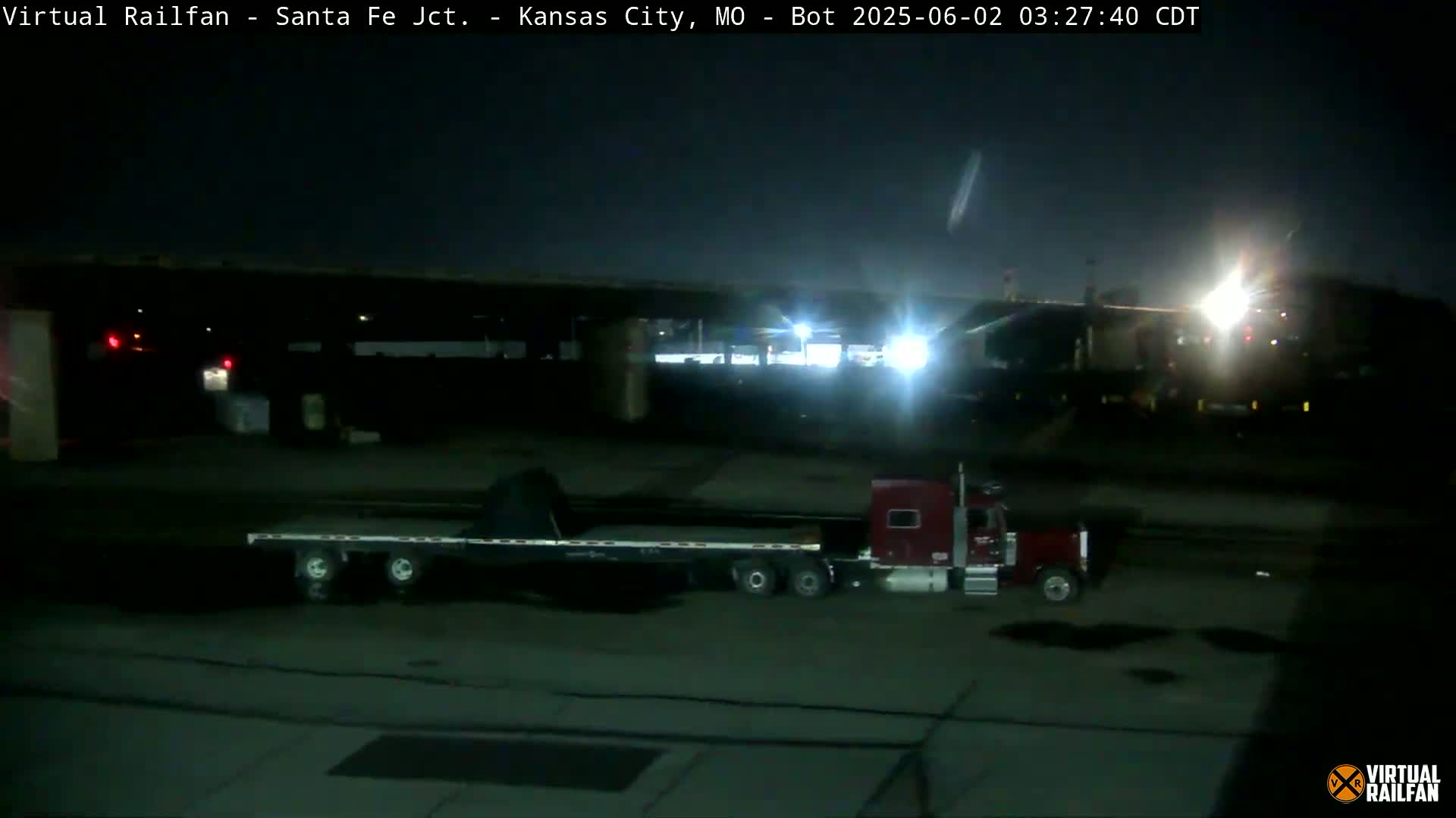 A red semi-truck and trailer are parked in a dark, nighttime lot under a bridge.