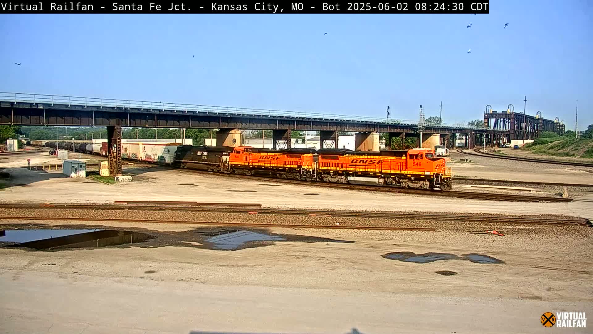 Under a clear blue sky, two orange locomotives lead a freight train across a rail yard, passing under a bridge.