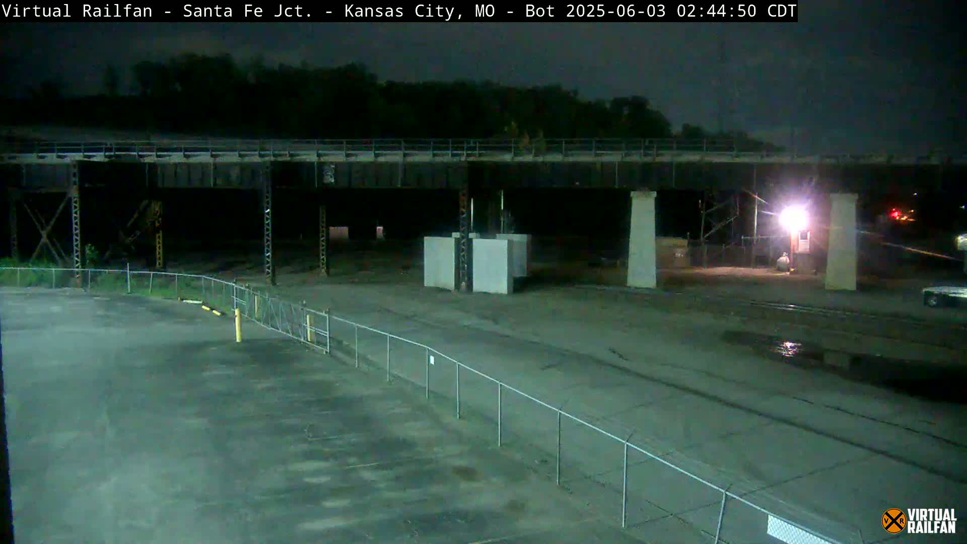 Here is a description of the image: Under a dark, nighttime sky, a large bridge spans over a mostly empty paved area enclosed by a chain-link fence.