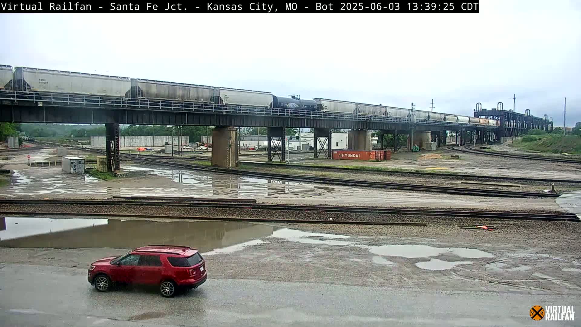 A long freight train crosses a railway bridge over a rain-soaked rail yard where a red SUV is parked.