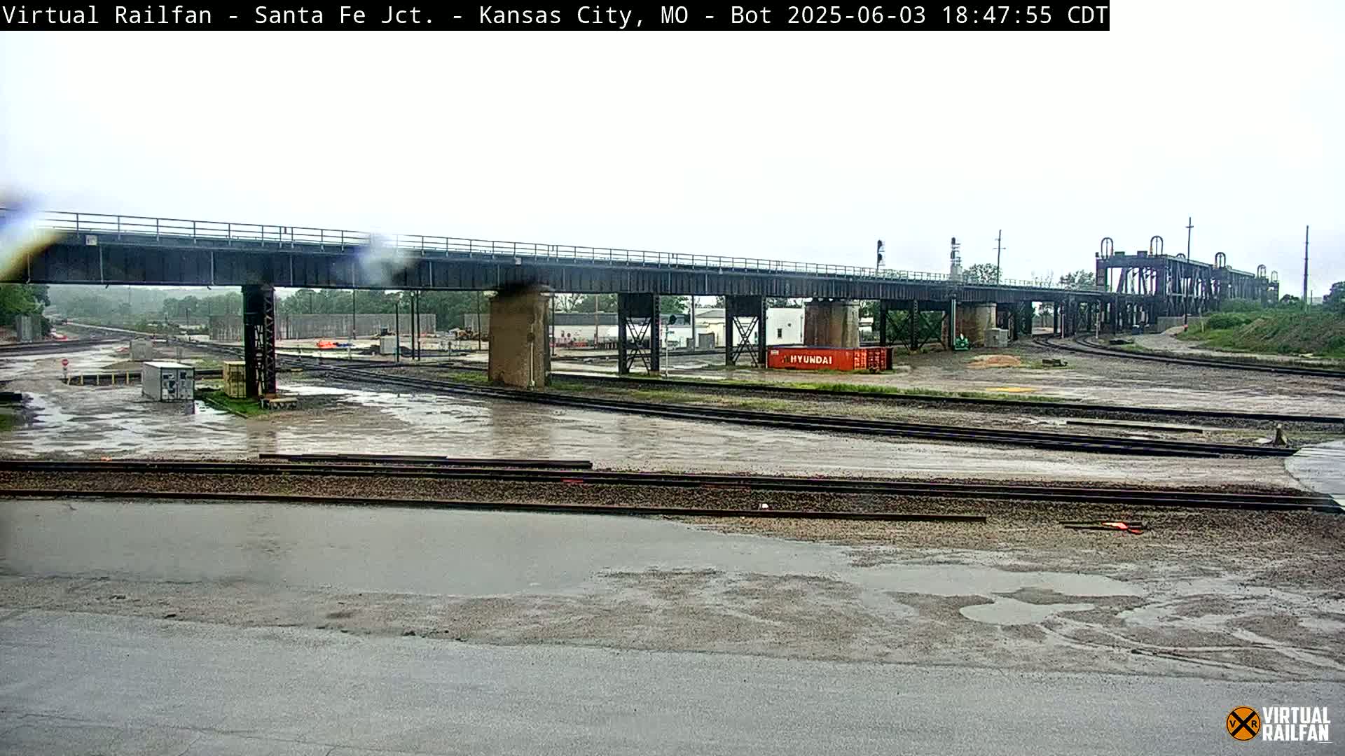 A railway yard with a bridge overhead, wet from recent rain, shows several sets of train tracks intersecting under an overcast sky.