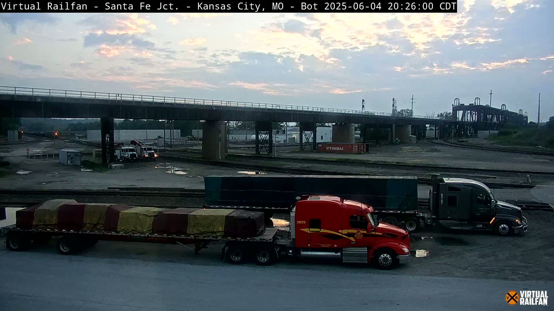 A twilight view of a railway yard with several semi-trucks and trailers parked near a bridge.