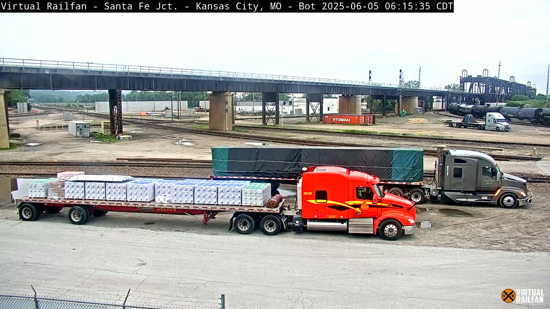 Two semi-trucks, one red and one gray, are parked in a rail yard under an overcast sky, with one truck carrying many packages on its trailer.
