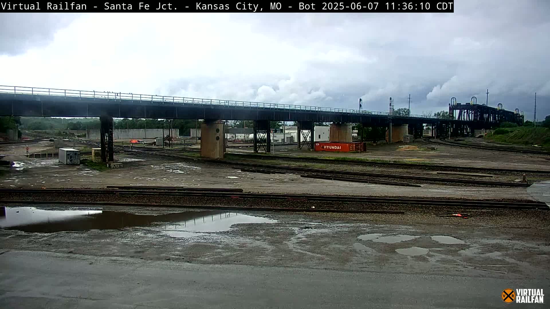 A railway yard with a large bridge overhead under an overcast sky, showing several puddles on the ground.
