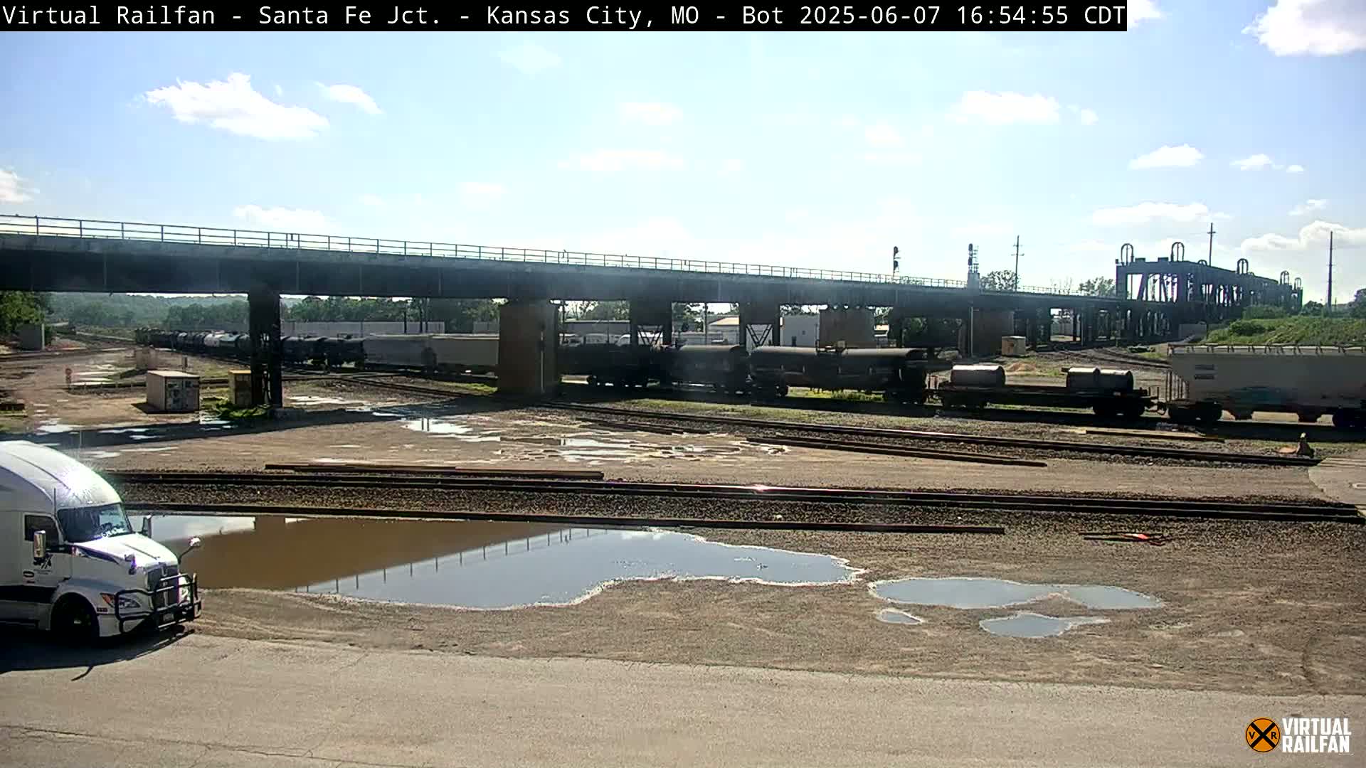 A white semi-truck is parked near railroad tracks in a yard with several train cars and a bridge overhead under a partly cloudy sky.