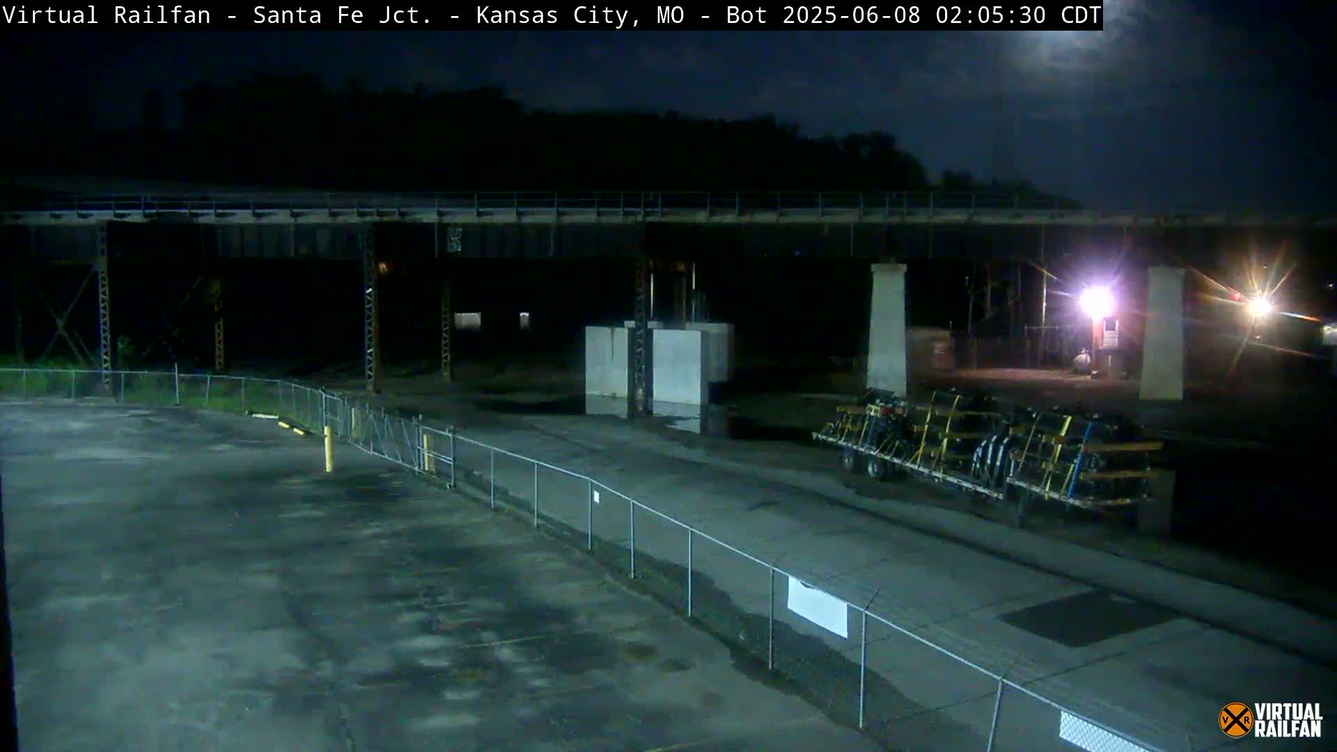 A nighttime view of a rail yard under a mostly clear sky shows a large bridge overhead, a flatbed trailer carrying cargo, and an empty paved area enclosed by a chain-link fence.