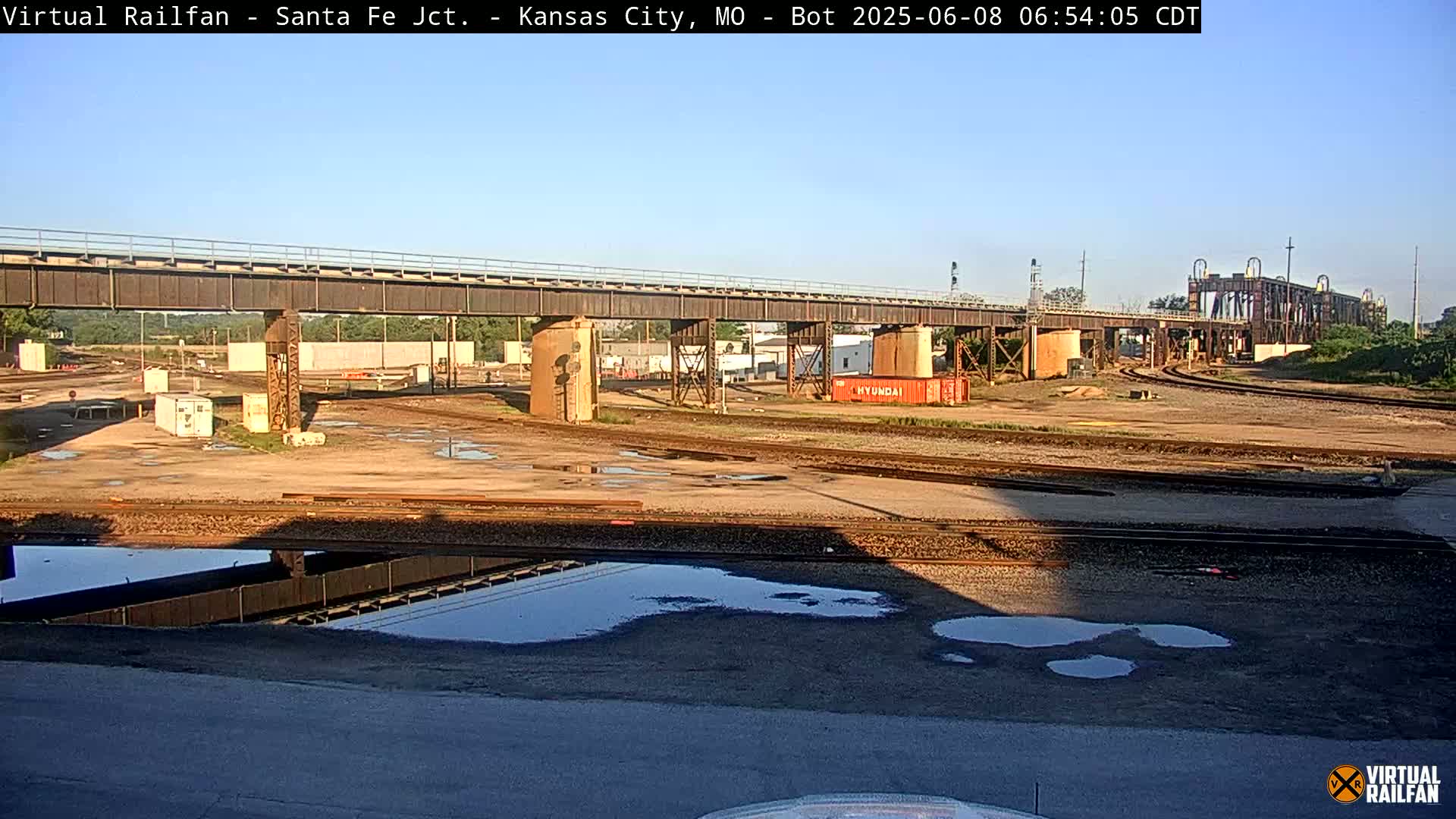 A large metal bridge spans over a railway yard with several puddles in a mostly sunny outdoor setting.