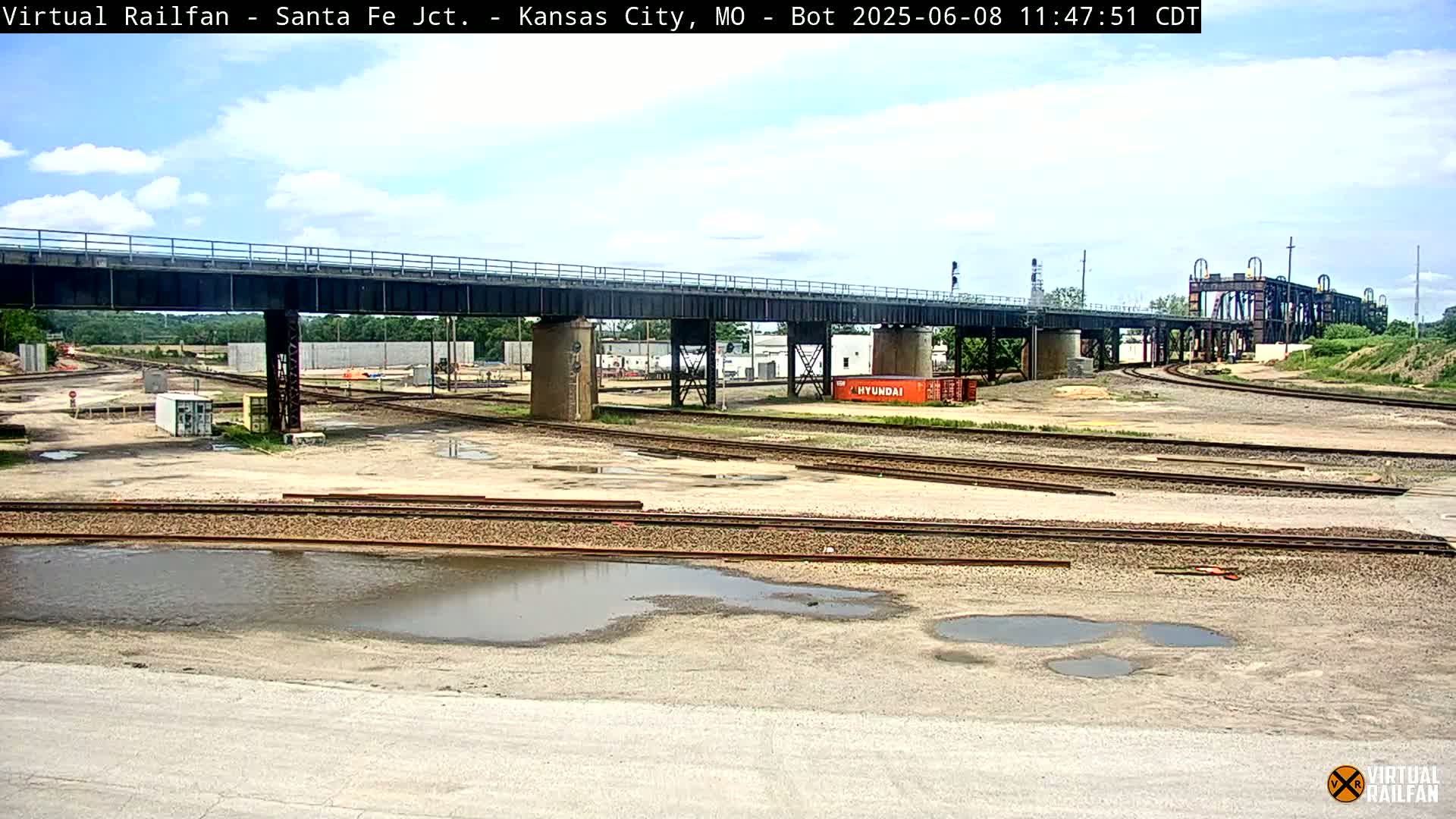 A railway yard with a bridge overhead, under a partly cloudy sky, showing several puddles on the ground.