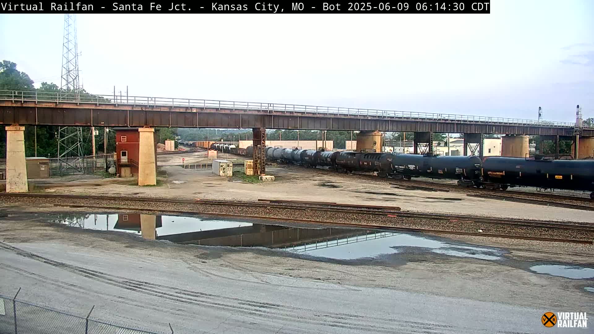 A long train of dark tanker cars sits under a bridge on a mostly overcast day, with puddles reflecting the scene in the foreground.