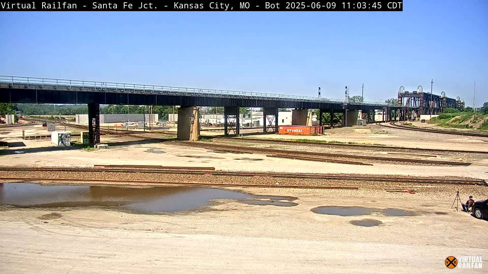 A railway yard under a bridge on a sunny day, with several puddles of water on the ground.