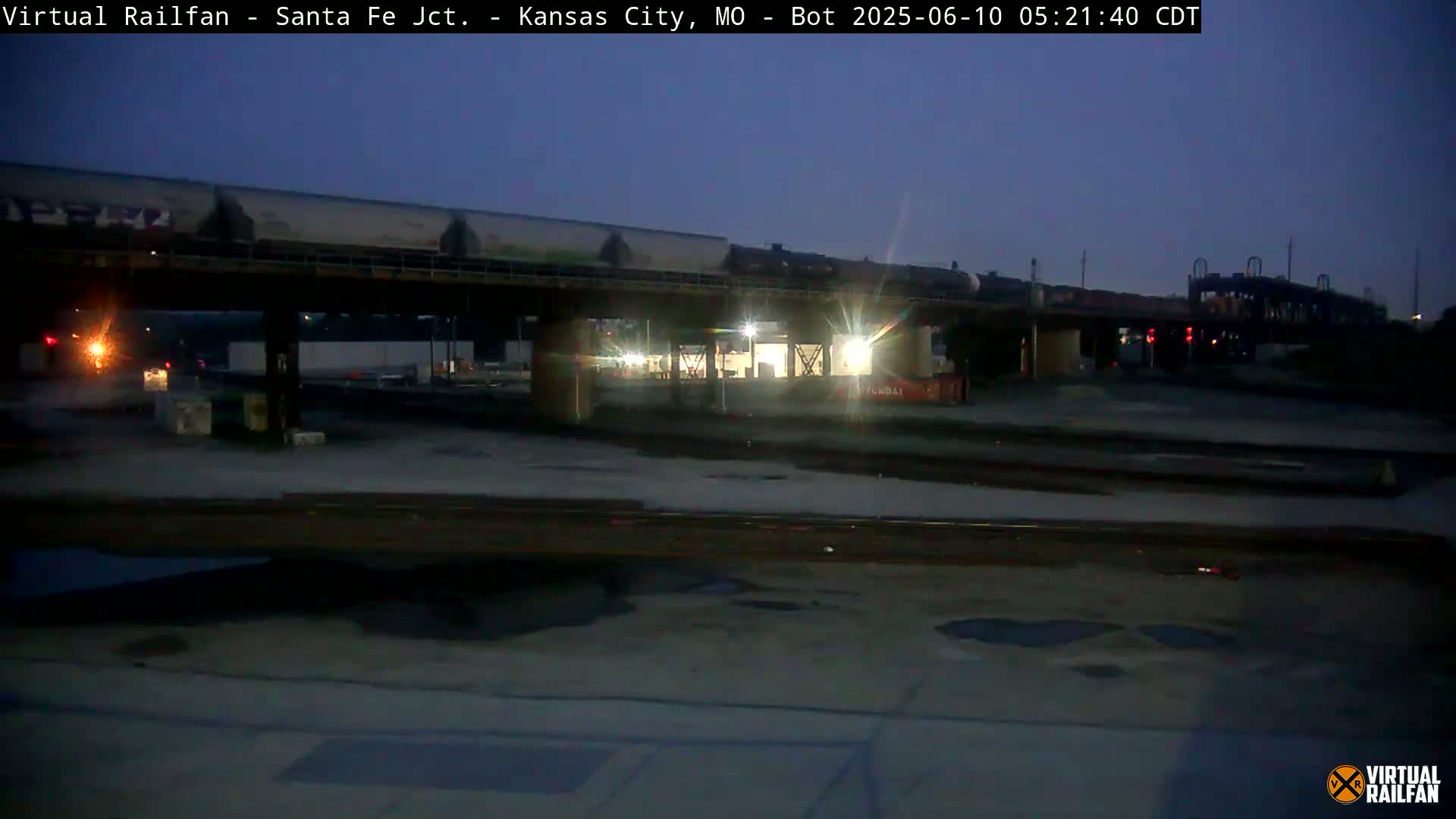 A long freight train passes under a bridge at twilight in a dimly lit railyard.