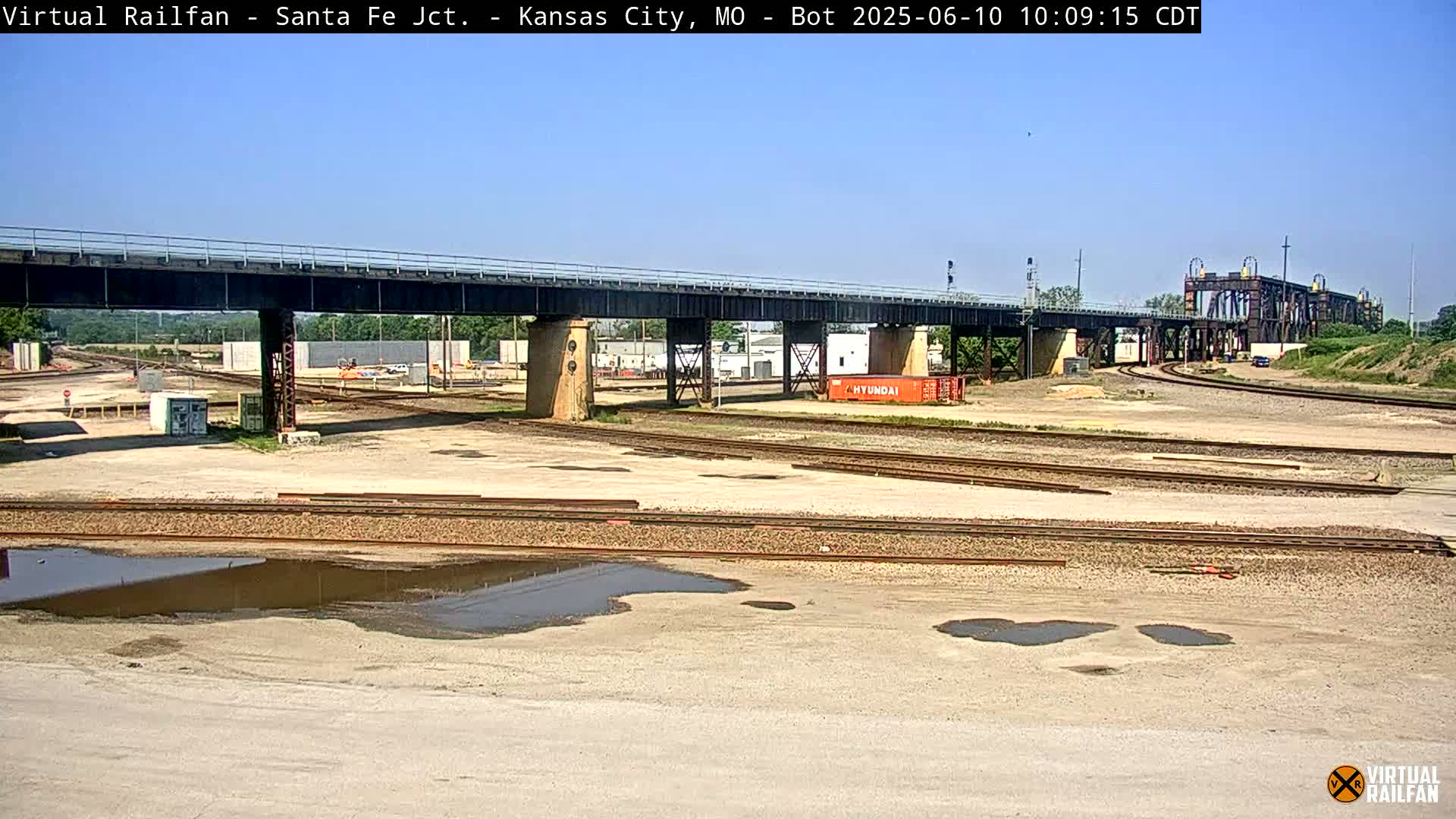 A railway yard with a large bridge under a clear blue sky, showing several sets of train tracks and puddles of water on the ground.