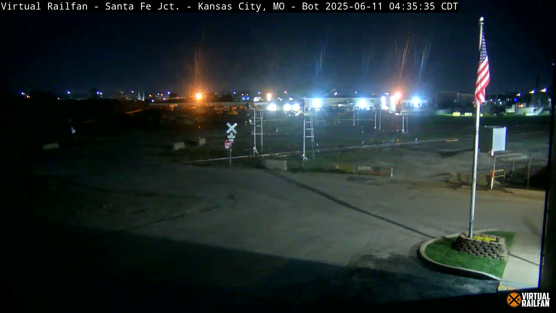 A nighttime view of a railroad crossing and surrounding area, with an American flag displayed on a pole, under clear skies.