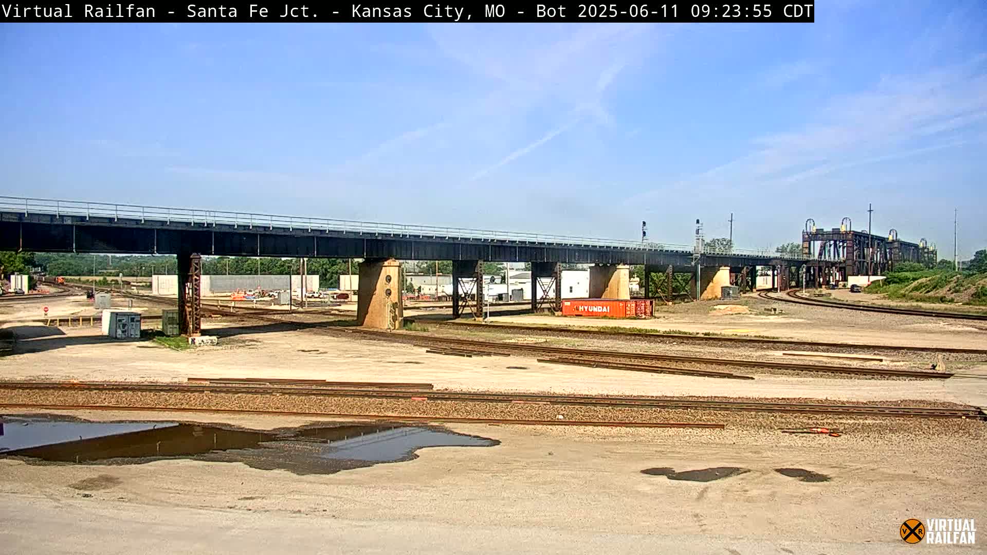 A railway yard with a large bridge overhead under a mostly clear, sunny sky.