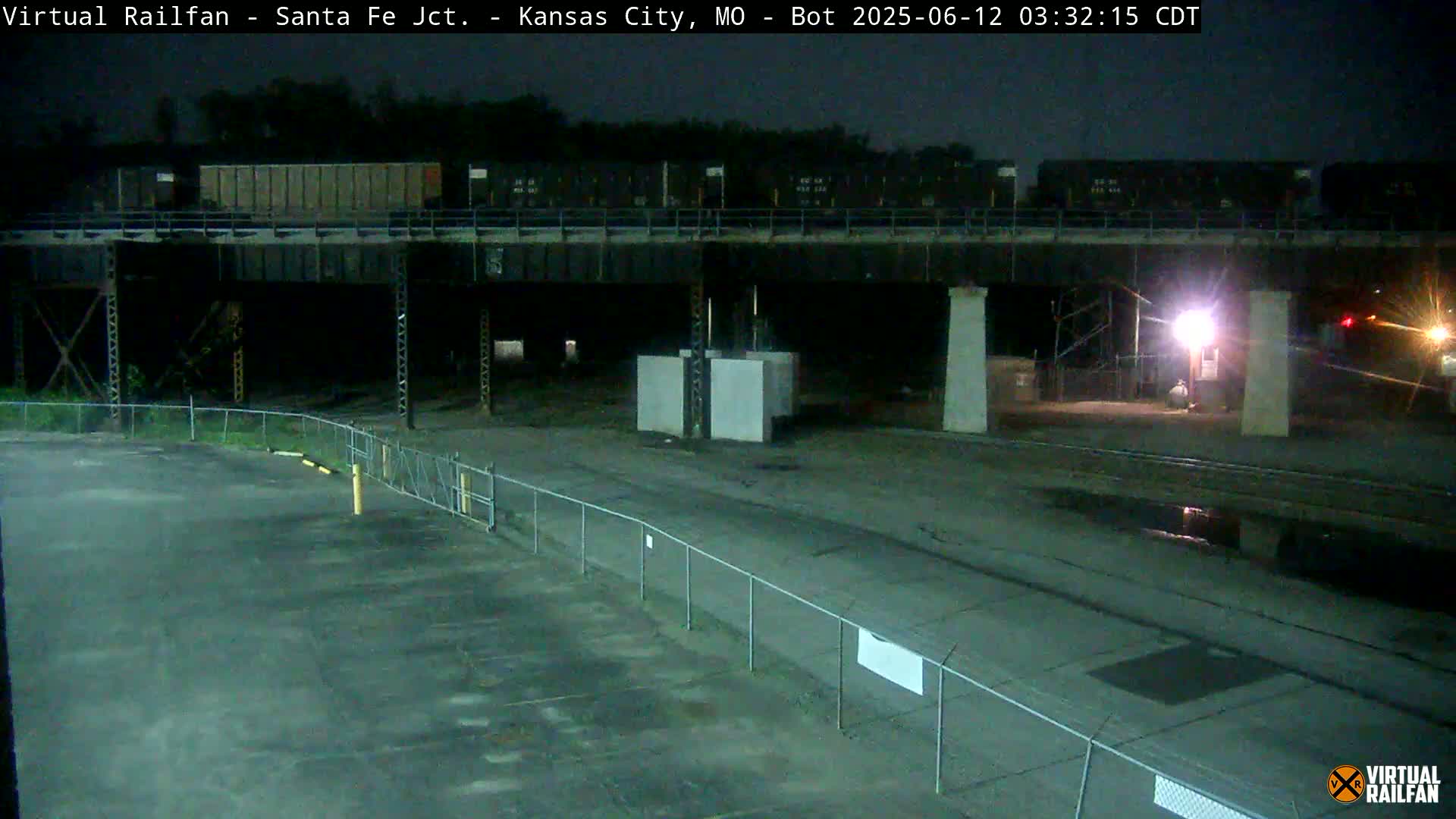 A nighttime view of a train on an overpass above a mostly empty asphalt lot enclosed by a chain link fence.