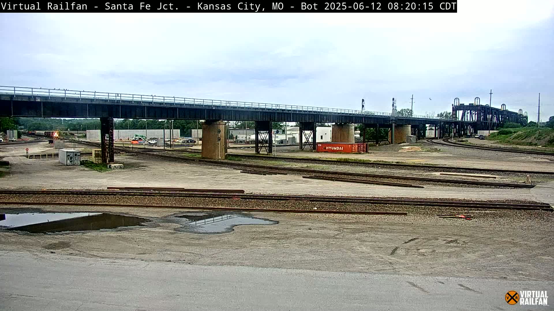 A railway yard with a large bridge overhead, under an overcast sky, shows multiple sets of train tracks, a few rail cars, and a puddle of water.