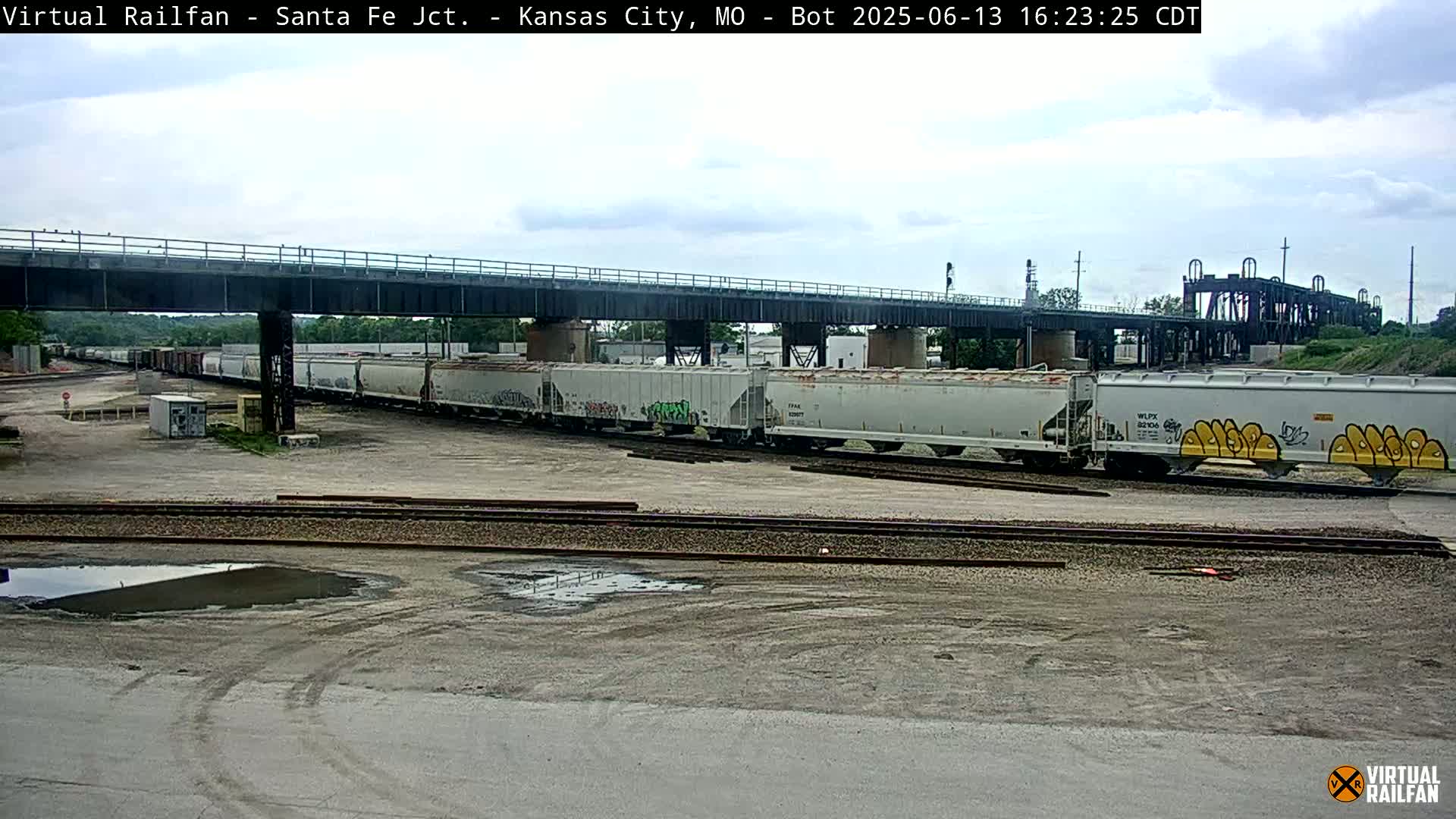 A long freight train sits under an overpass on a partly cloudy day.
