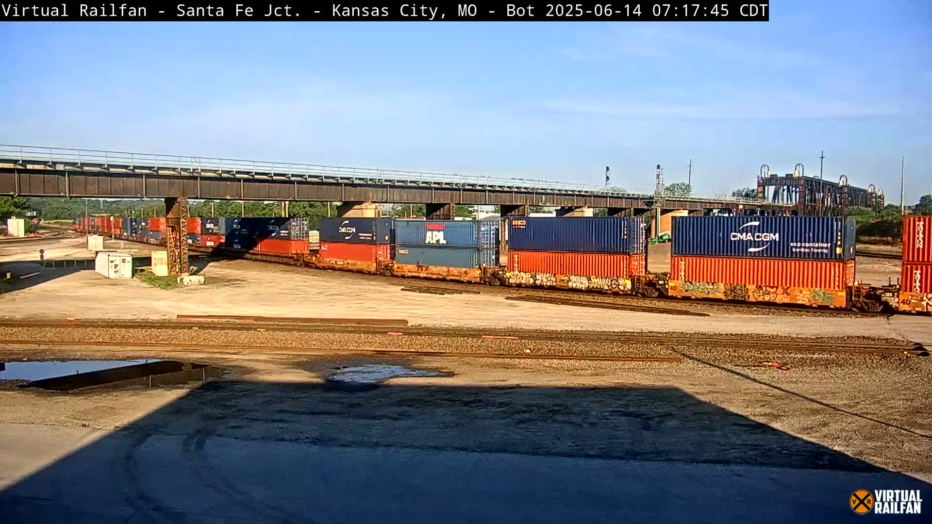 Under a clear sky, a long train carrying numerous colorful shipping containers sits parked beneath a railway bridge.