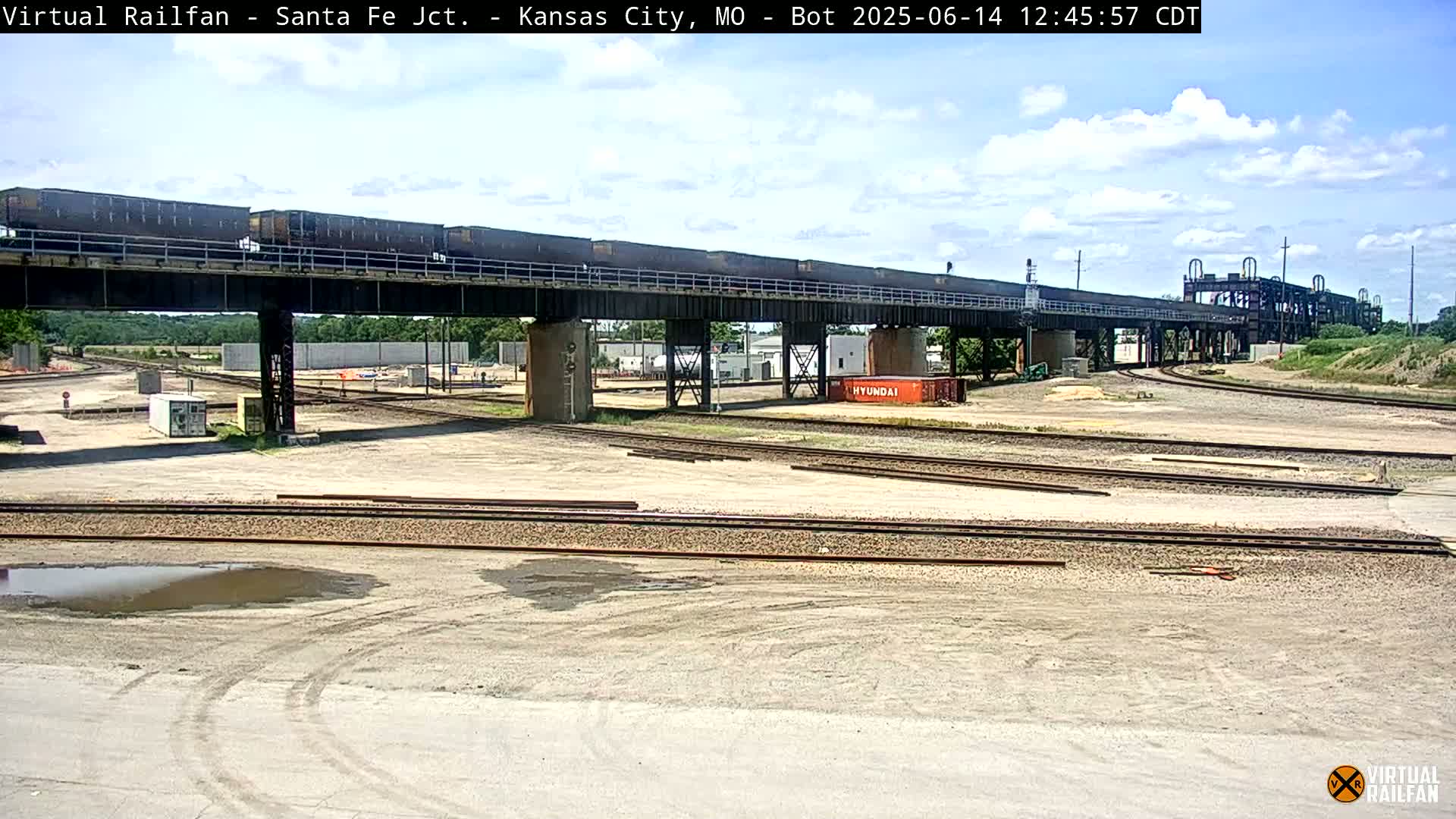 A train carrying many dark-colored boxcars crosses a railway bridge under a partly cloudy sky.