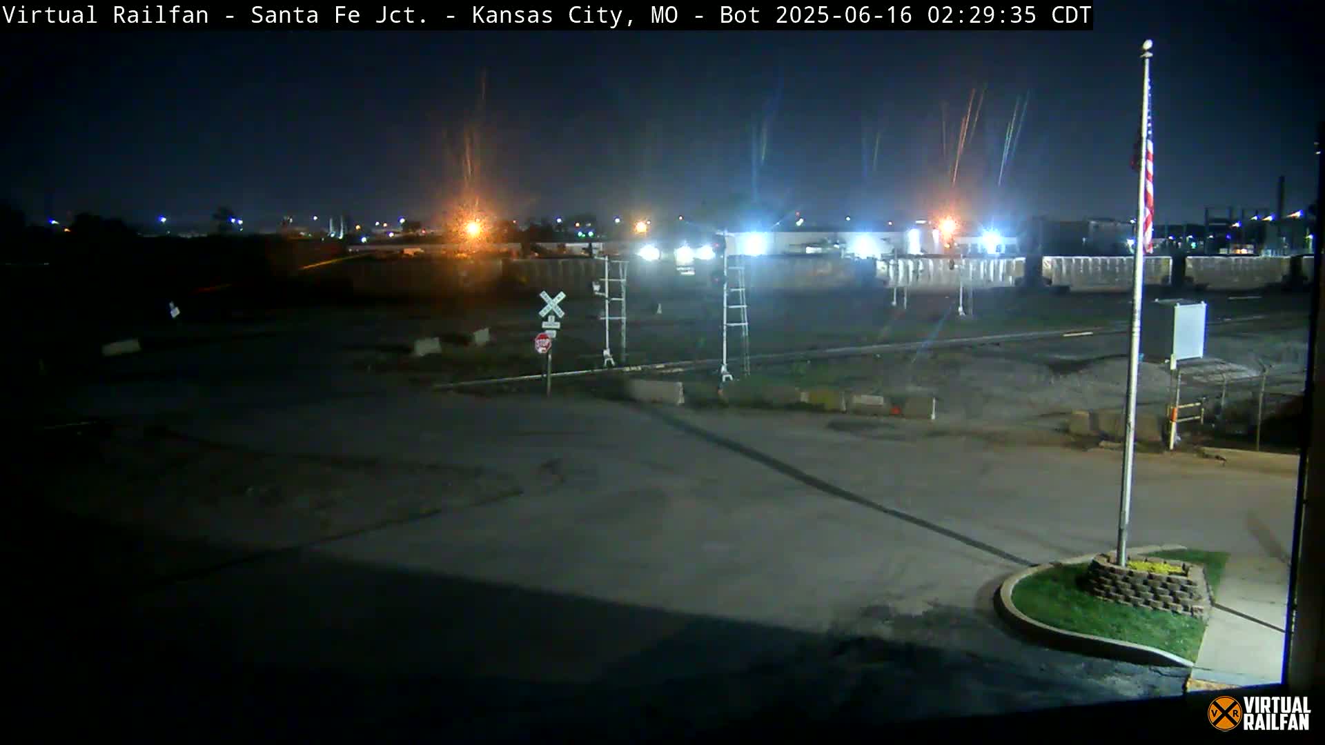 A nighttime view of a railroad crossing near a collection of railcars and industrial buildings under a clear, dark sky.
