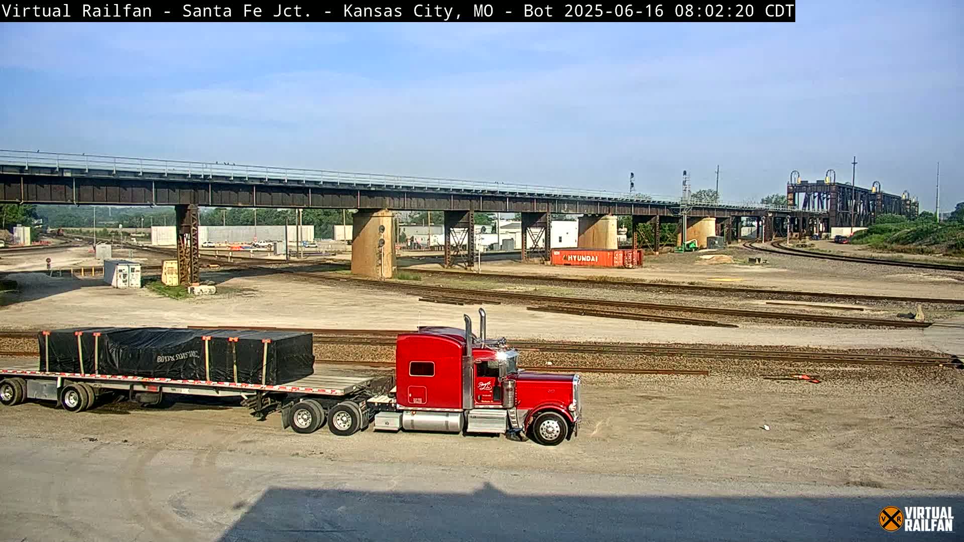A red semi-truck hauling a flatbed trailer sits in a rail yard under a mostly clear sky, with a large bridge and several sets of railroad tracks visible in the background.