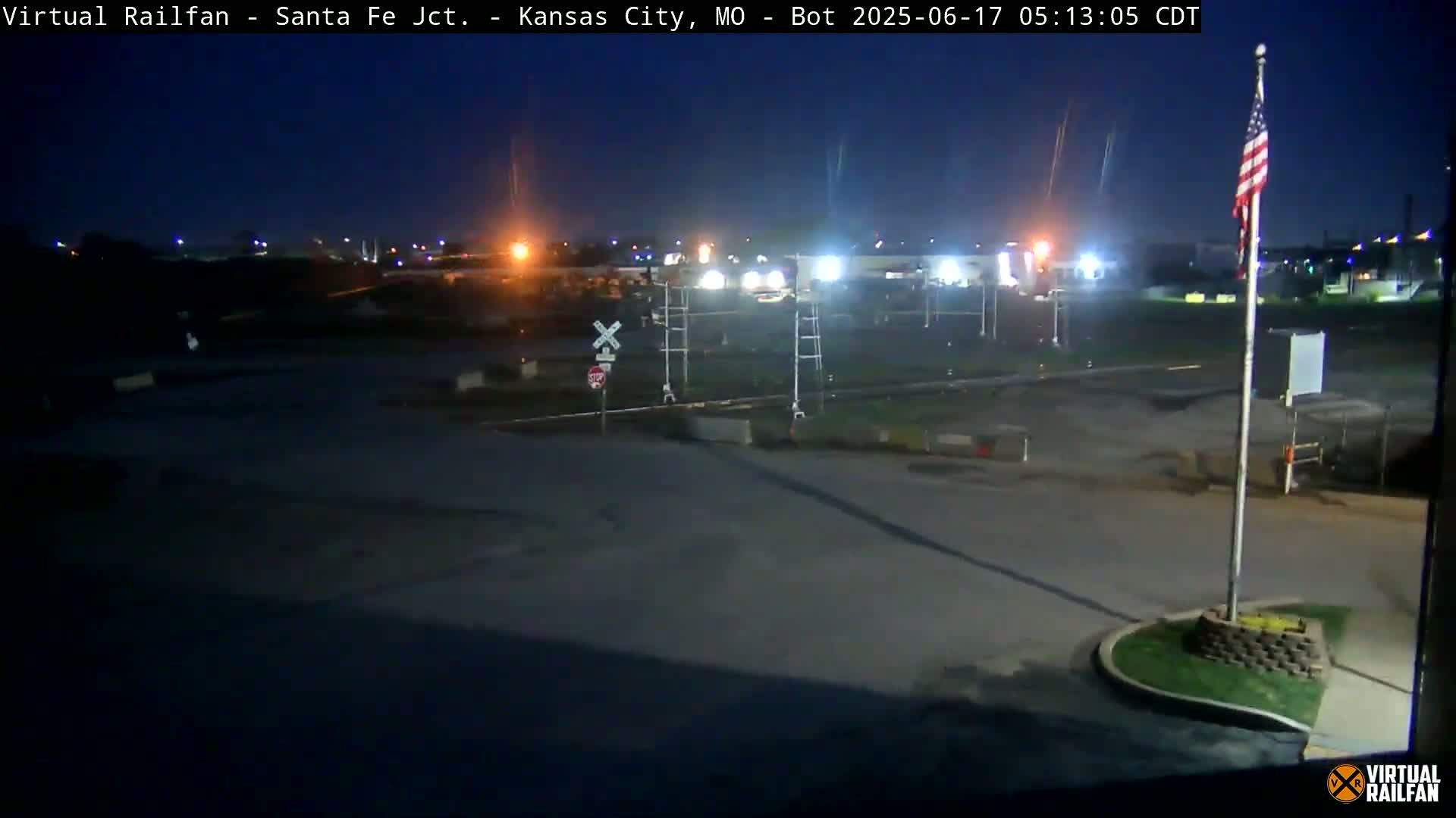 A nighttime view of a railroad crossing and surrounding area, with a flagpole bearing an American flag and dim, distant lights visible under a dark sky.
