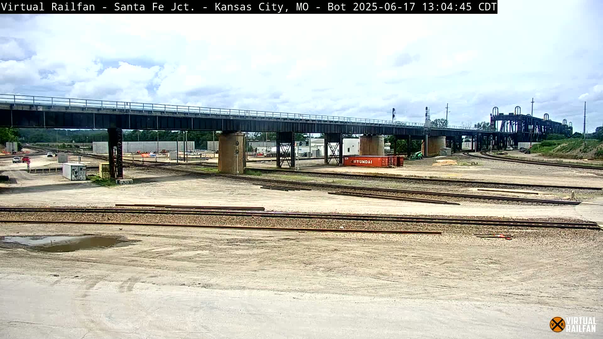 A mostly sunny day shows a railway yard with multiple train tracks, a large bridge over the yard, and a single orange shipping container.
