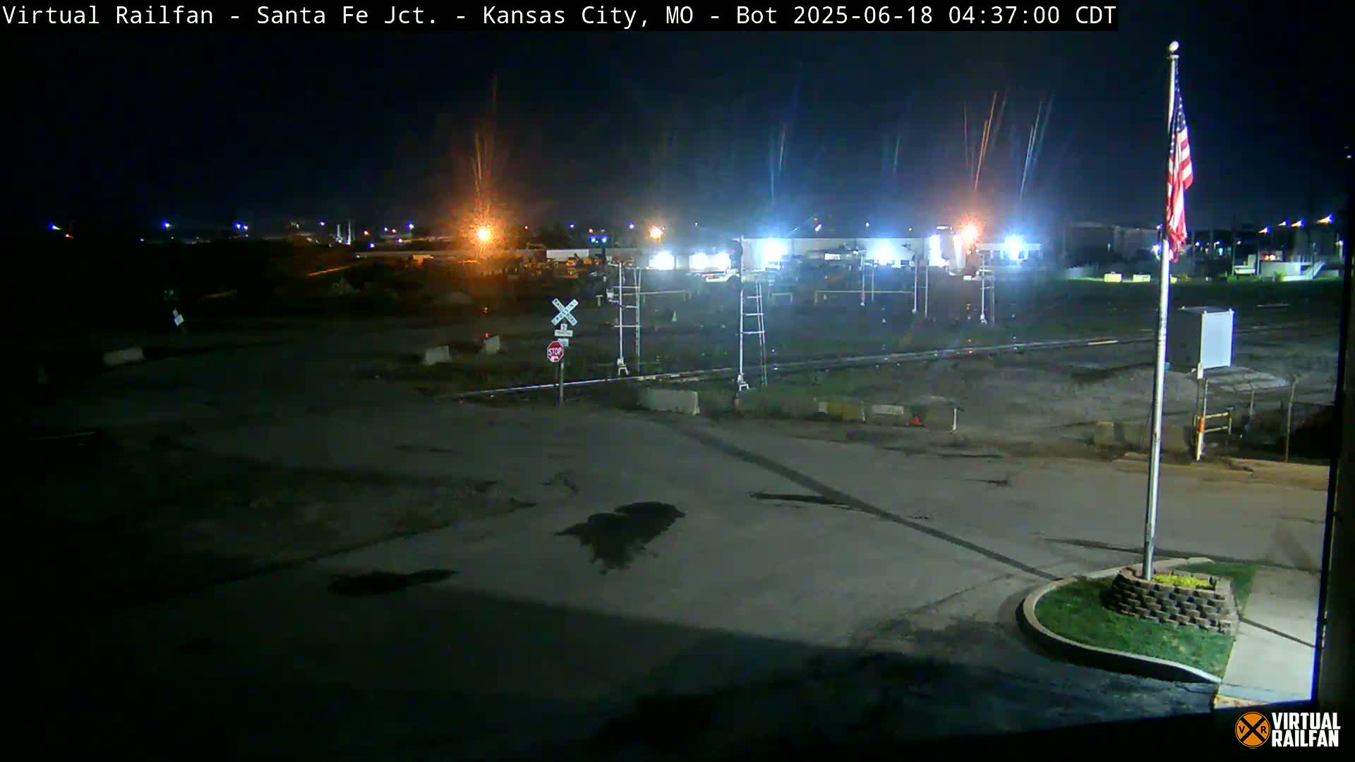 A nighttime view of a mostly empty rail yard with a railroad crossing and an American flag on a pole, under a clear sky.