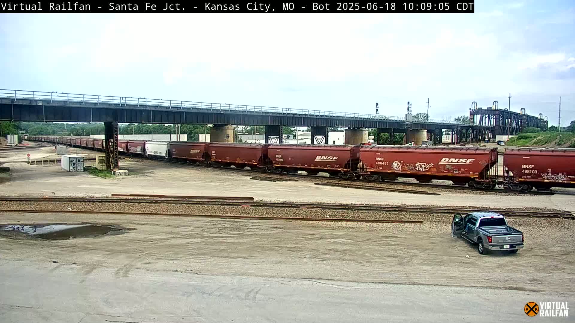 A long freight train sits under a bridge on a partly sunny day, with a pickup truck parked nearby in a mostly empty rail yard.