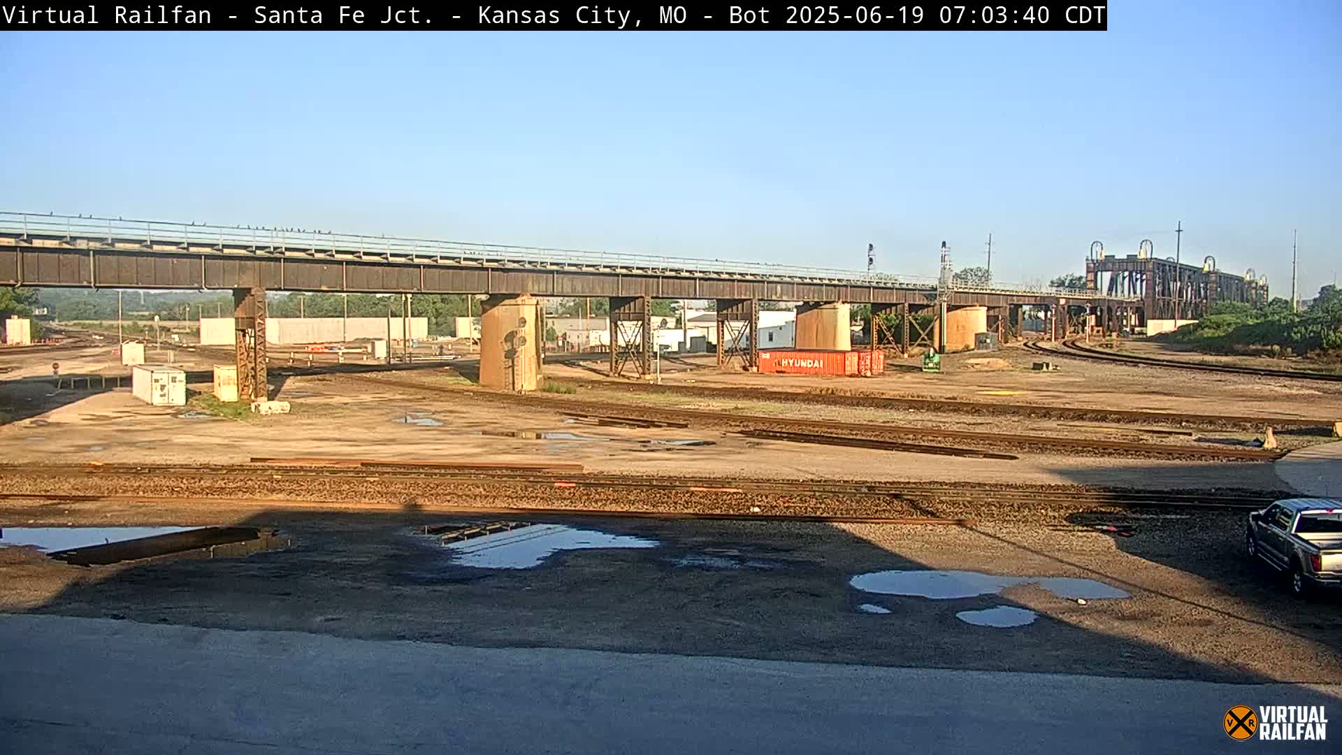 Under a clear sky, a large railroad yard with several train tracks, a bridge, and a few containers is visible, with some puddles on the ground.