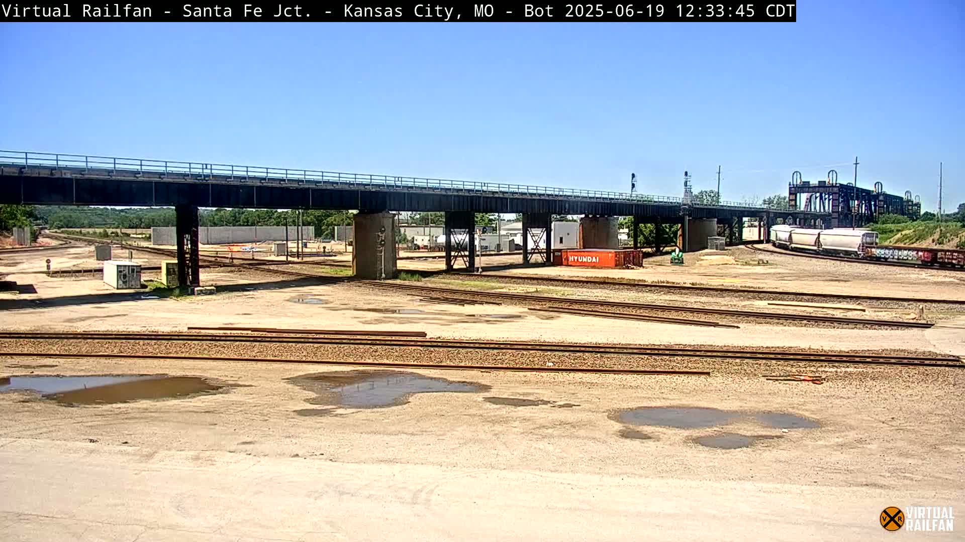 A railway yard with multiple train tracks, a bridge overhead, and a few train cars under a clear, sunny sky.